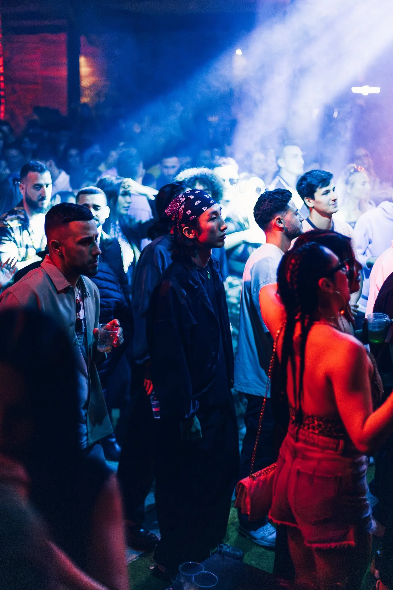 People attending a concert or party under colorful stage lighting, some holding drinks, with a diverse crowd in a dark, lively atmosphere.