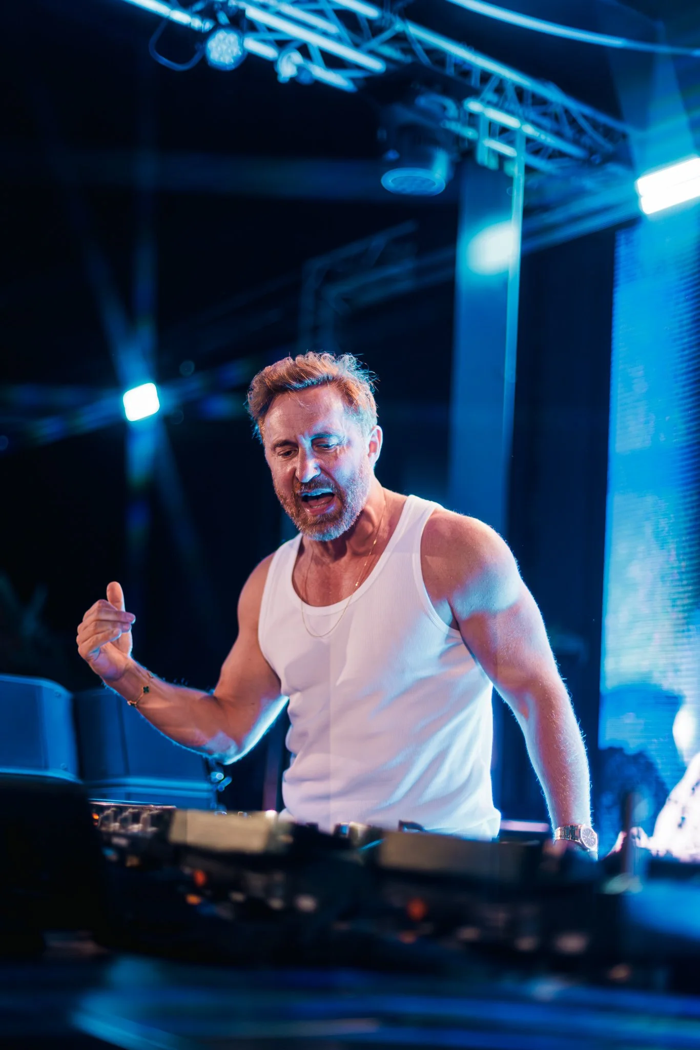 A man with gray hair and a beard wearing a white tank top DJing at a club or event, with lighting and equipment visible in the background.