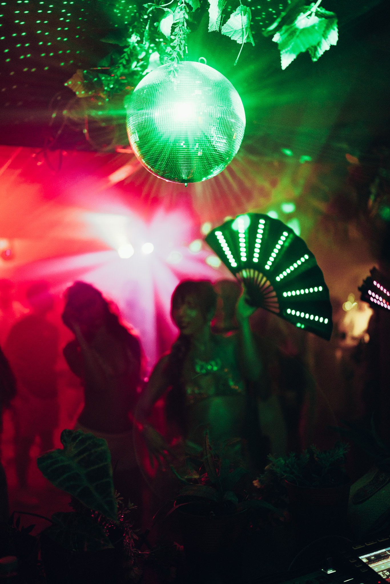 A green disco ball hanging from the ceiling with green and red lights shining around. The silhouettes of several people are visible, some with plants or leaves around them, suggesting a party or dance event with a tropical or jungle theme.