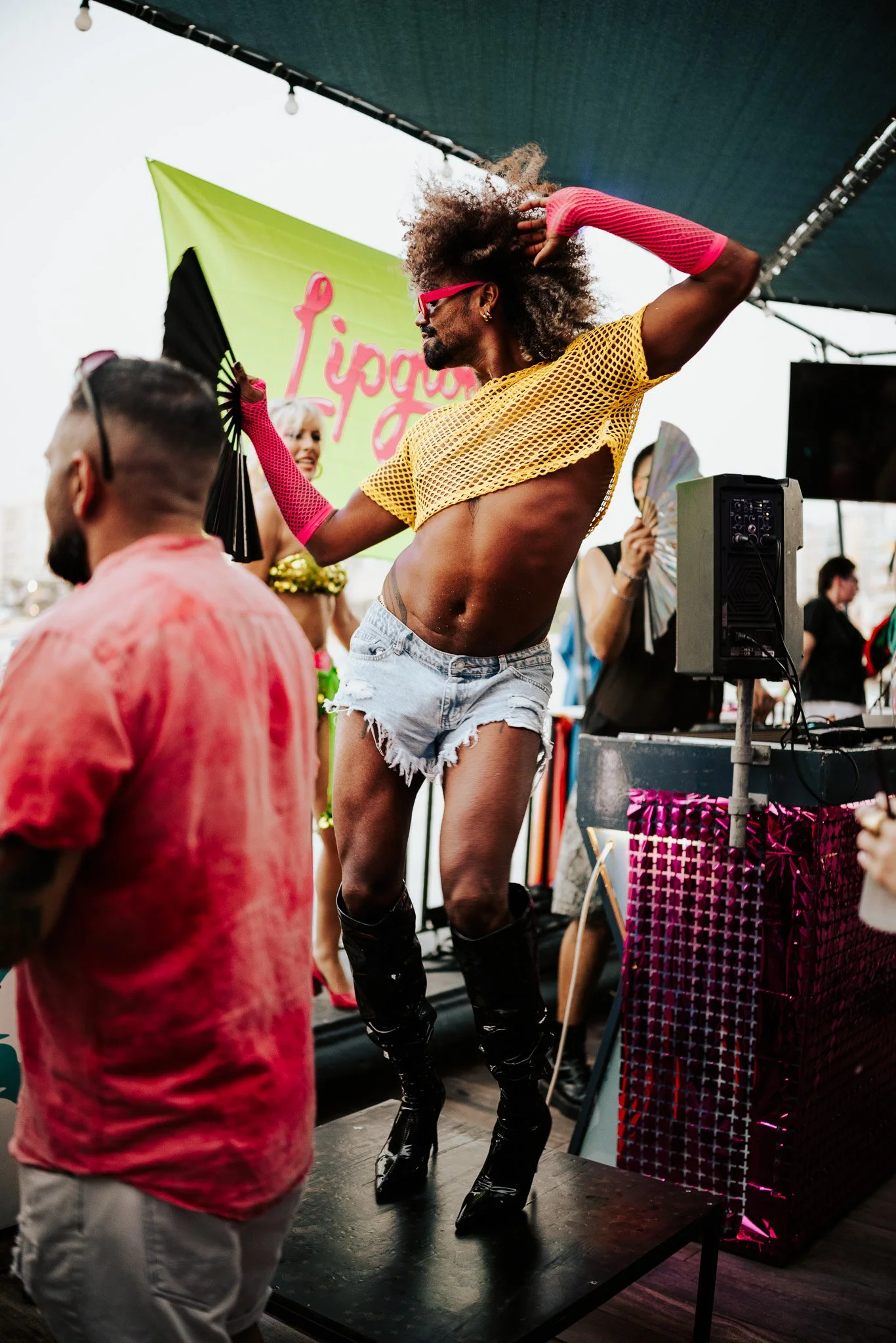 A person dancing at an outdoor event with a lime green backdrop behind them that has pink writing on it. They are wearing a yellow fishnet top, distressed denim shorts, pink fishnet gloves, and high black boots. Several people are visible around them