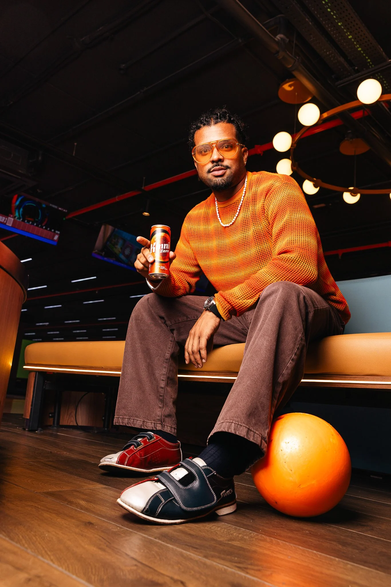 guy at the bowling alley, holding a kinnie, wearing an orange sweater, orange bowling ball next to his feet, winter shoot