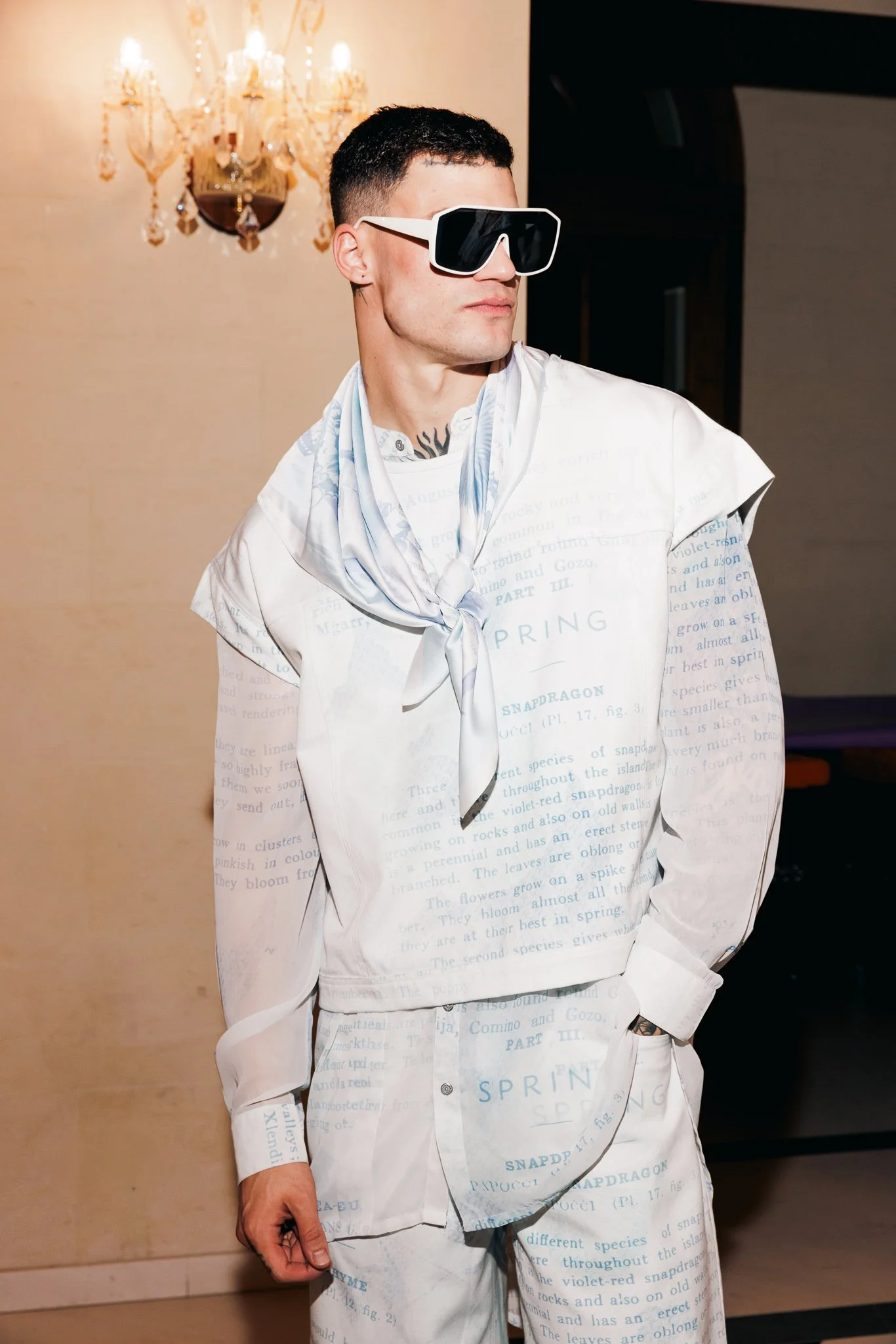 A young man wearing large black and white sunglasses, a white shirt with printed text, and a light-colored scarf, standing indoors in front of a beige wall and a chandelier with multiple lights.