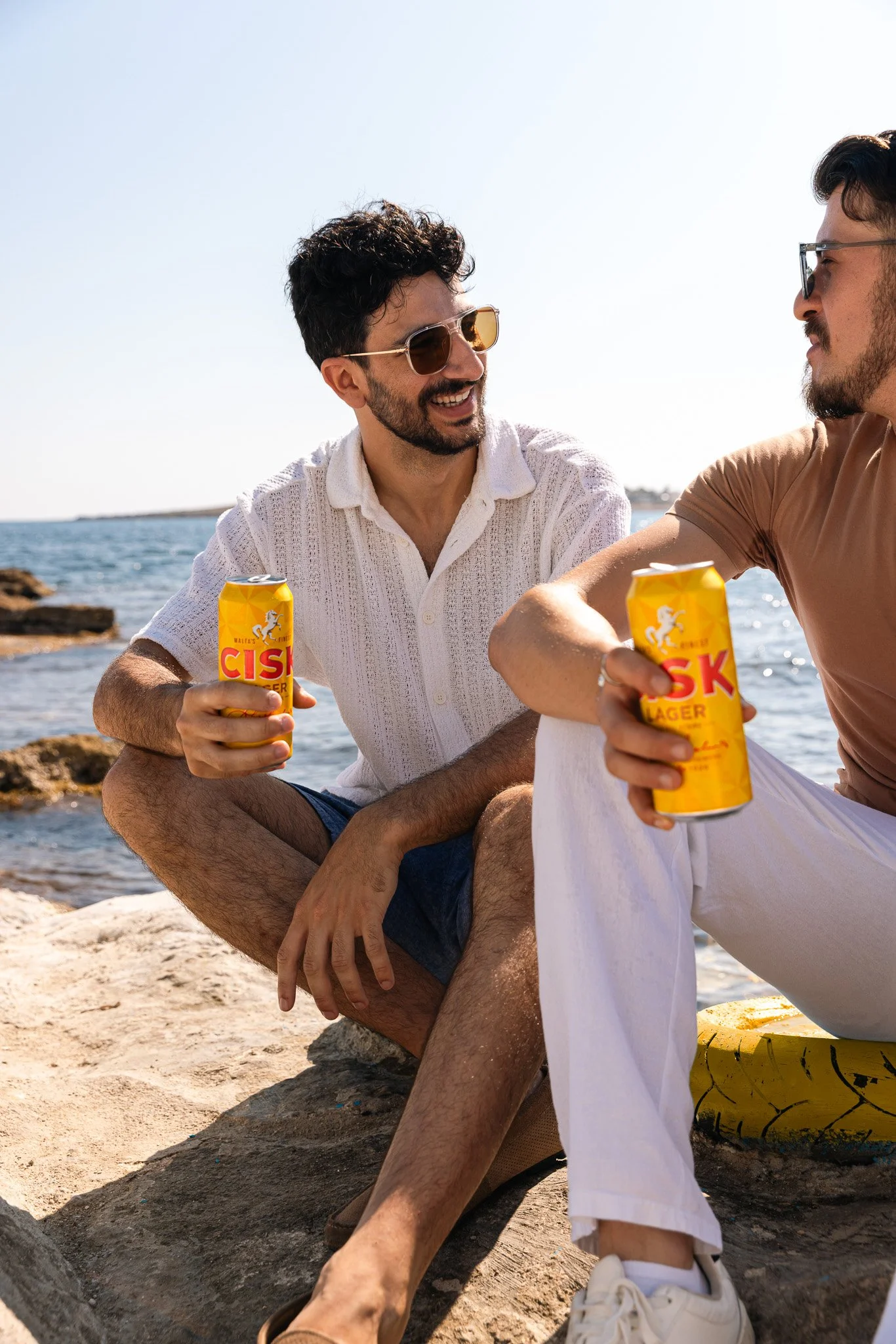 two men having a conversation by the beach, holding a cisk lager can