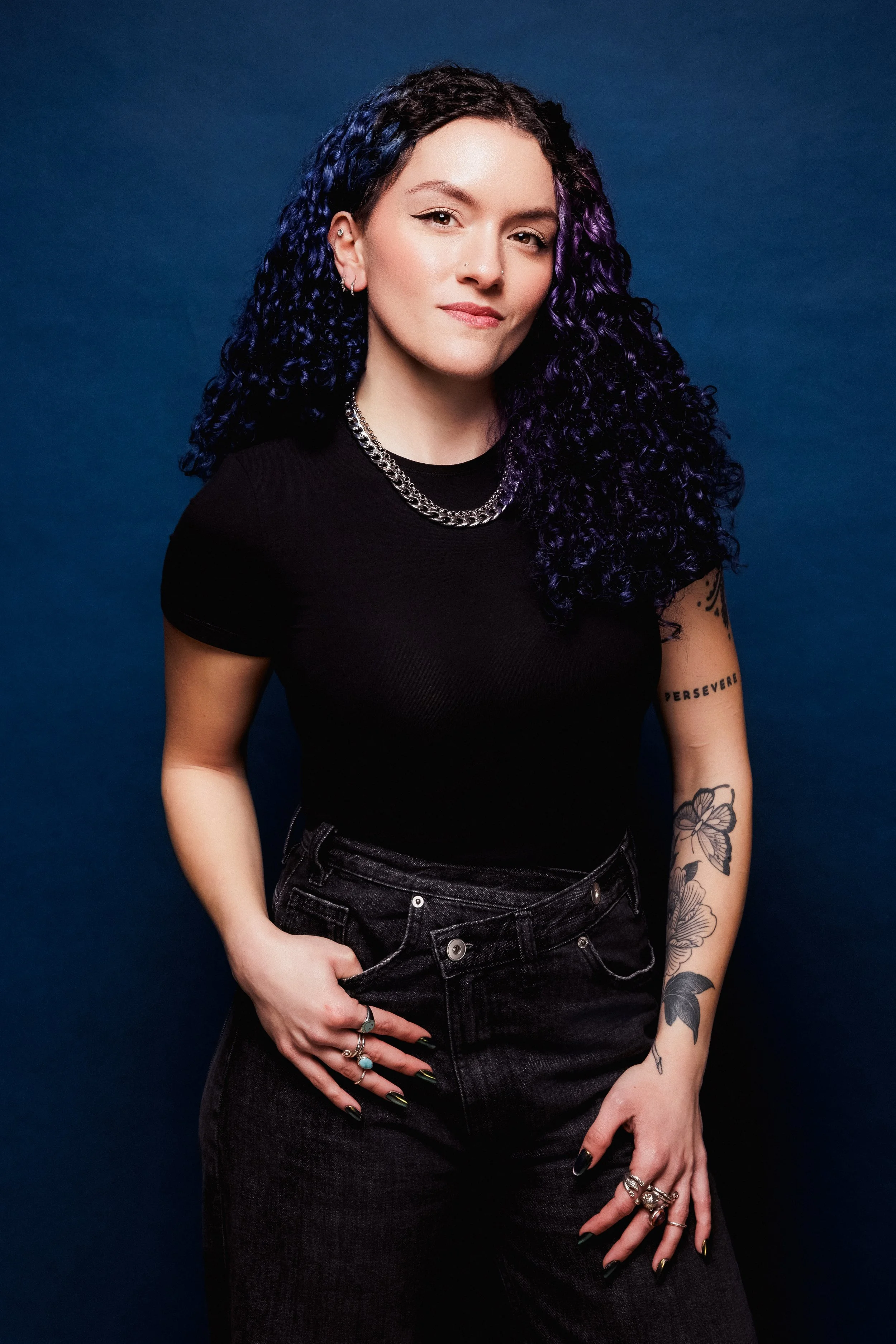 A woman with curly black and purple hair, wearing a black t-shirt and dark jeans, adorned with jewelry, and tattoos on her arms, stands against a dark blue background.