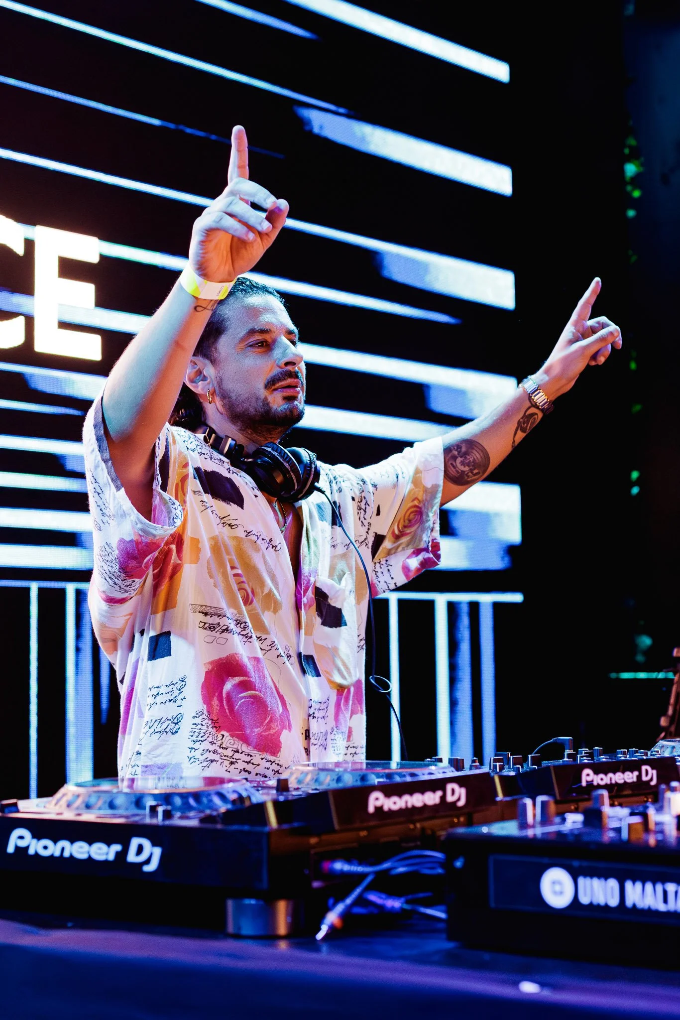 A DJ performing at a music event, standing behind a Pioneer DJ equipment, with both hands raised, one pointing upward and the other signaling peace, in front of a background of horizontal blue LED lights.