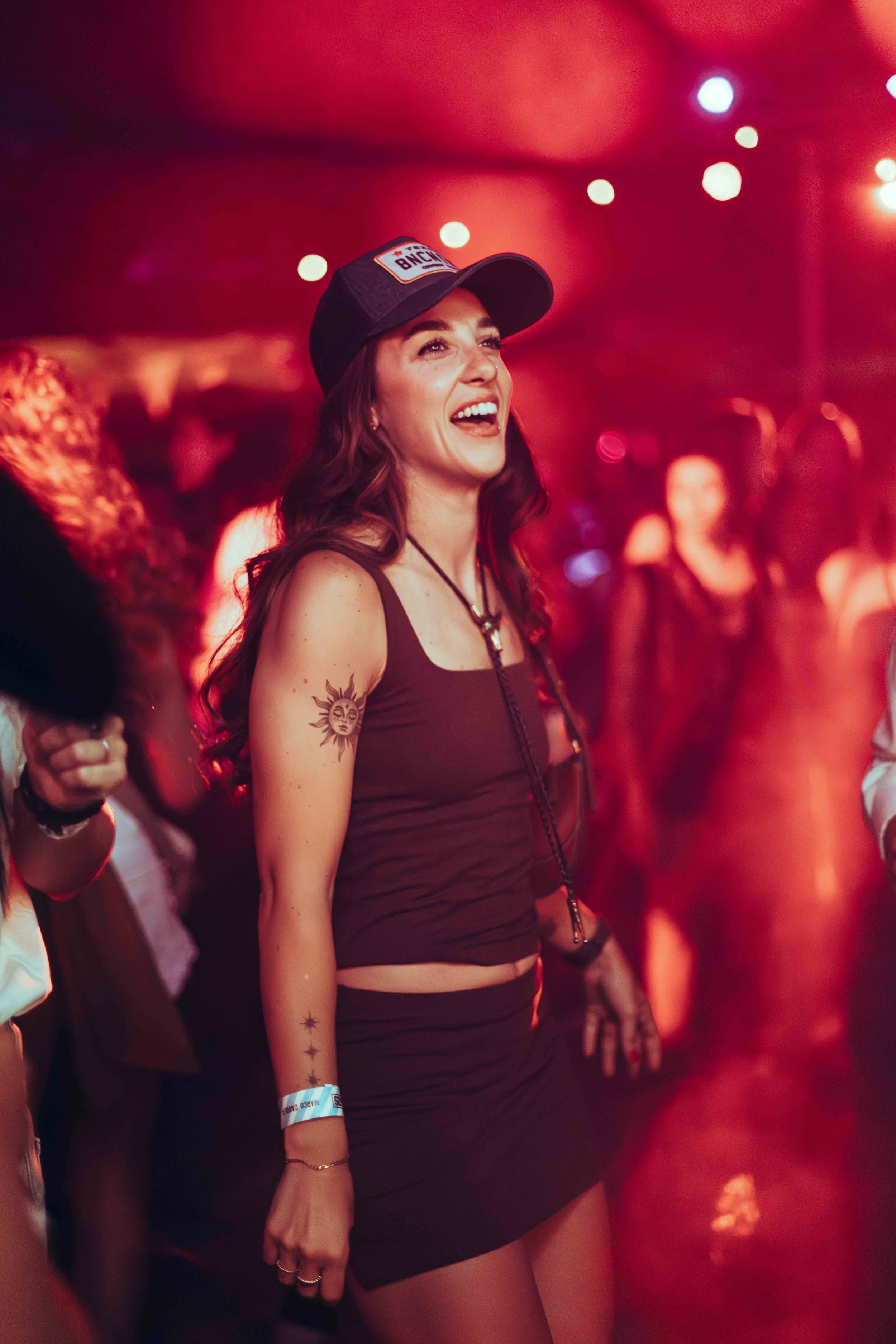 A woman with long brown hair, tattoos on her arm, and a wide smile at a music or dance event, wearing a black cap, black tank top, and black shorts, with colorful wristbands on her wrist. Bright, colorful lights and other people are in the background
