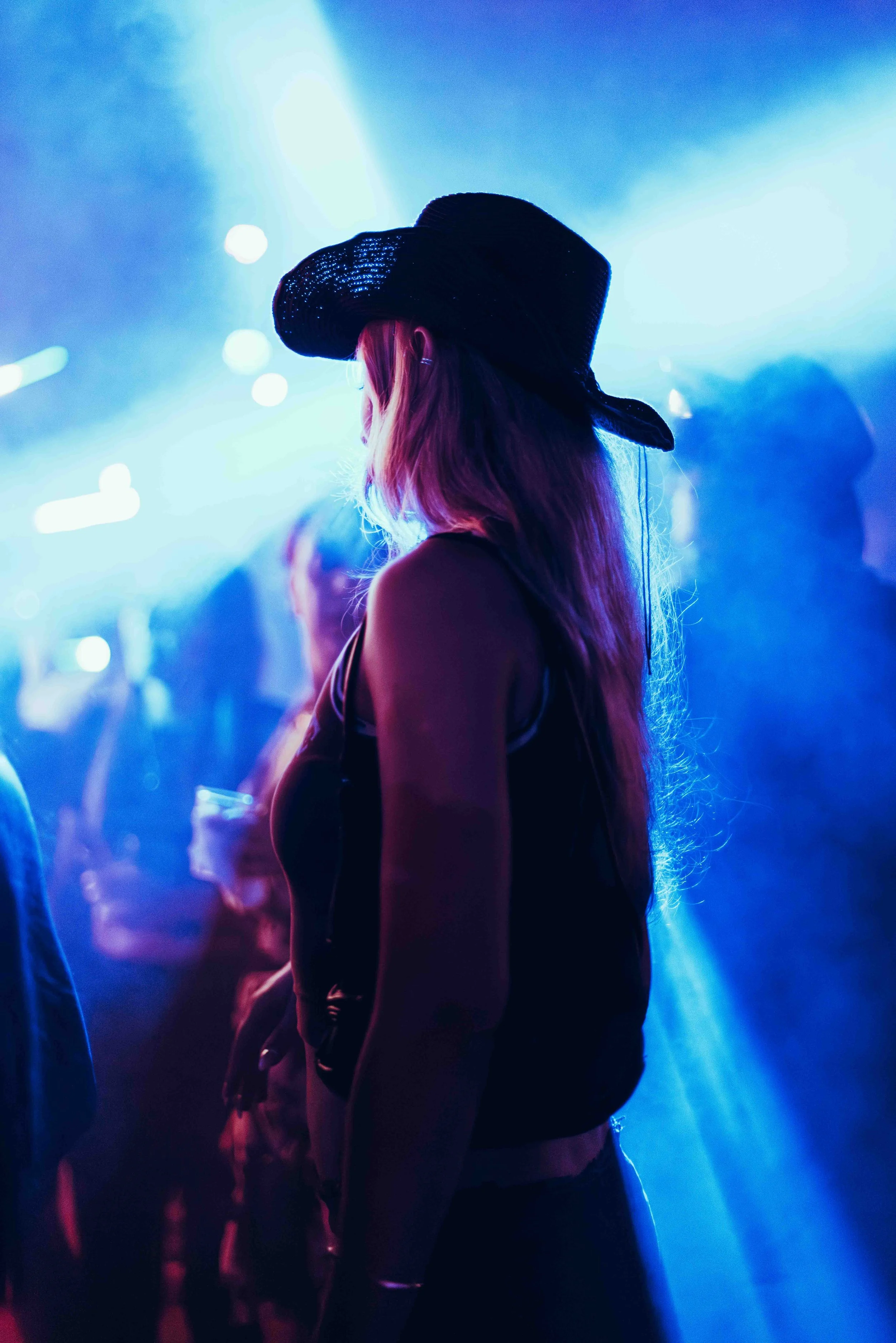 A woman with long hair wearing a large black hat, standing in a dimly lit venue with blue and purple neon lighting.