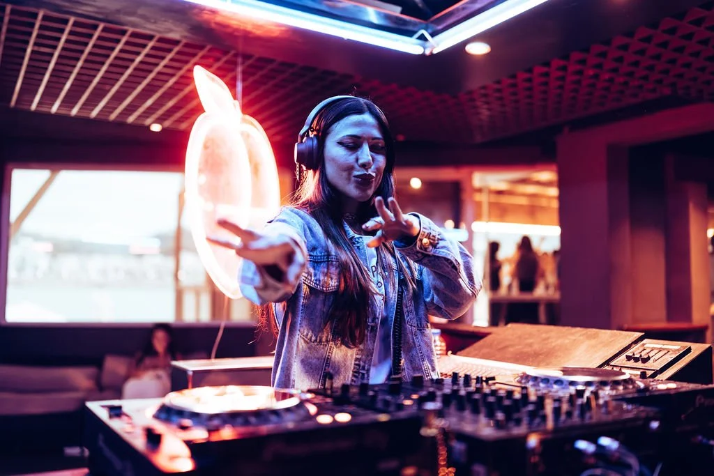 A woman DJ wearing headphones is performing in a dimly lit club with neon lighting, standing behind a DJ booth with turntables.