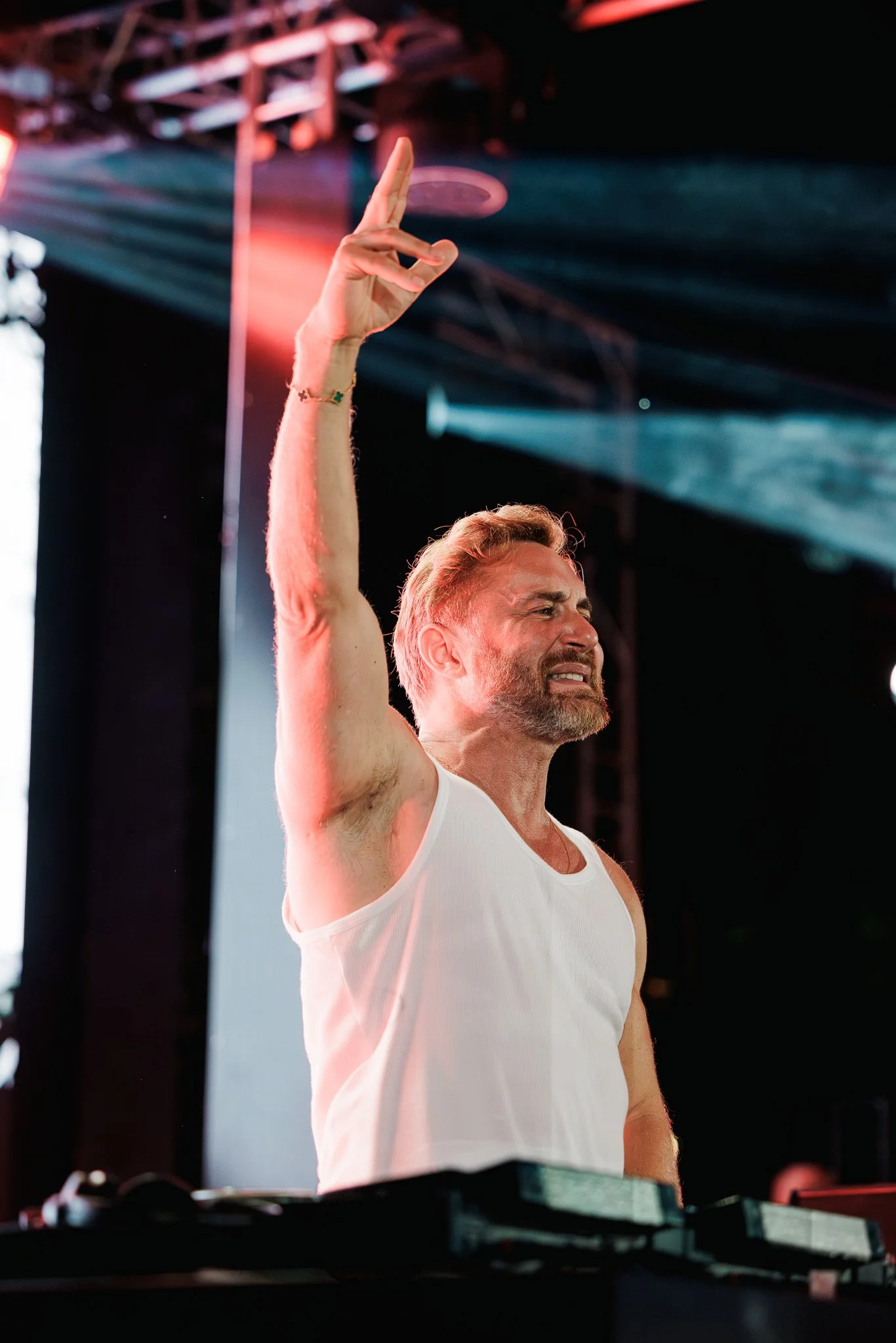 Man with a beard and short hair raising his right arm with a peace sign gesture at a music event or party, wearing a white sleeveless shirt, with stage lighting and blurred equipment in the background.