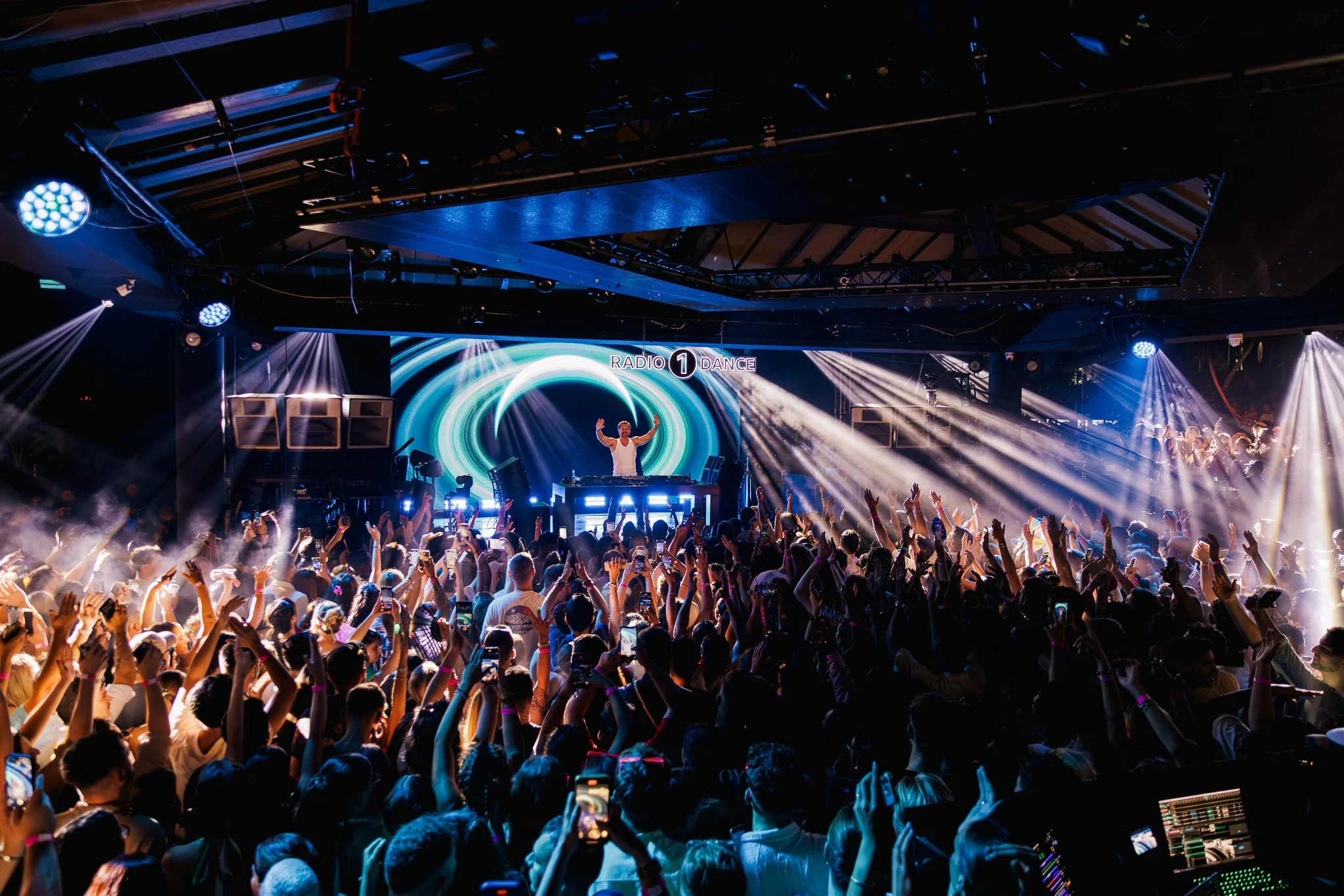 Crowd of people dancing and raising their hands at a concert with a DJ performing on stage under colorful lights and a large screen displaying 'Radio 1 Dance'.