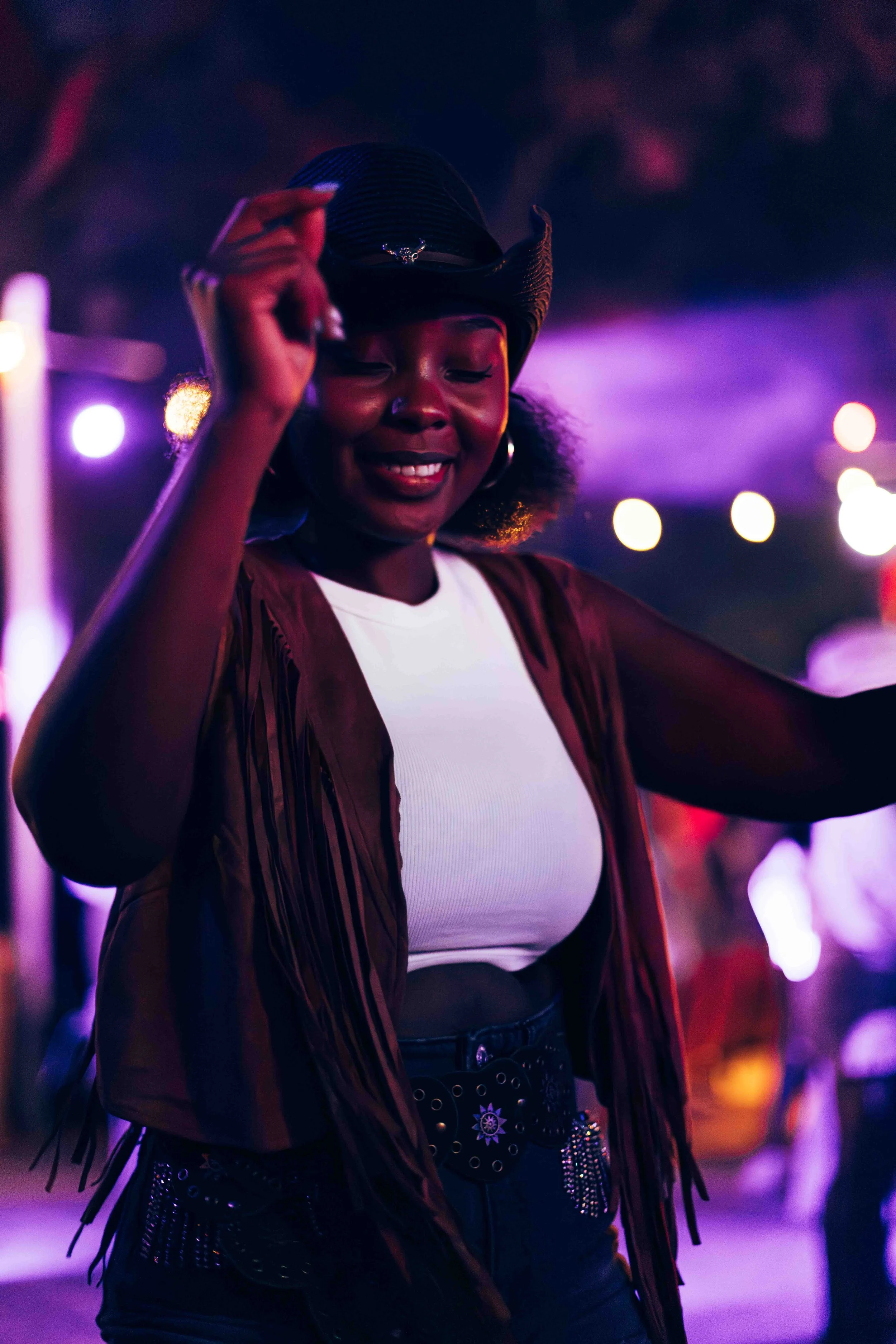 Young woman dancing at night with purple and orange city lights in background, wearing a black headwrap, red jacket, white shirt, and decorated belt.