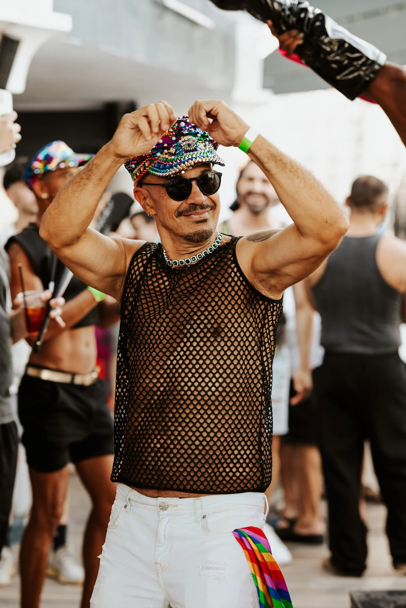 A person wearing a glittery, colorful hat, sunglasses, a black fishnet top, and white shorts, holding a rainbow pride flag, at a pride event with other participants in the background.