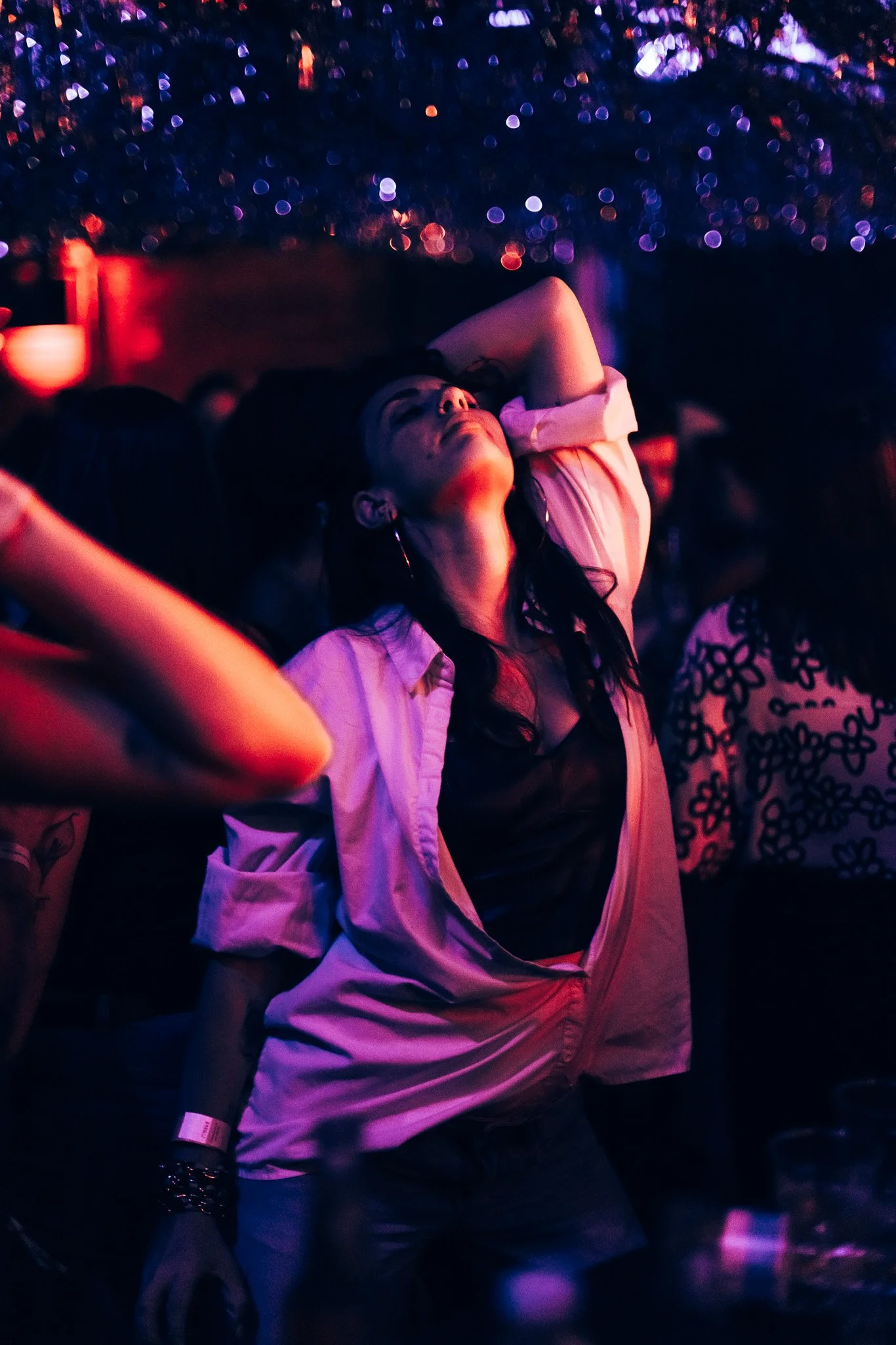 A woman with dark hair and earrings is in a dark room with colorful, bokeh-style lights in the background. She has one arm raised with her hand resting on her head, and she appears to be enjoying herself.
