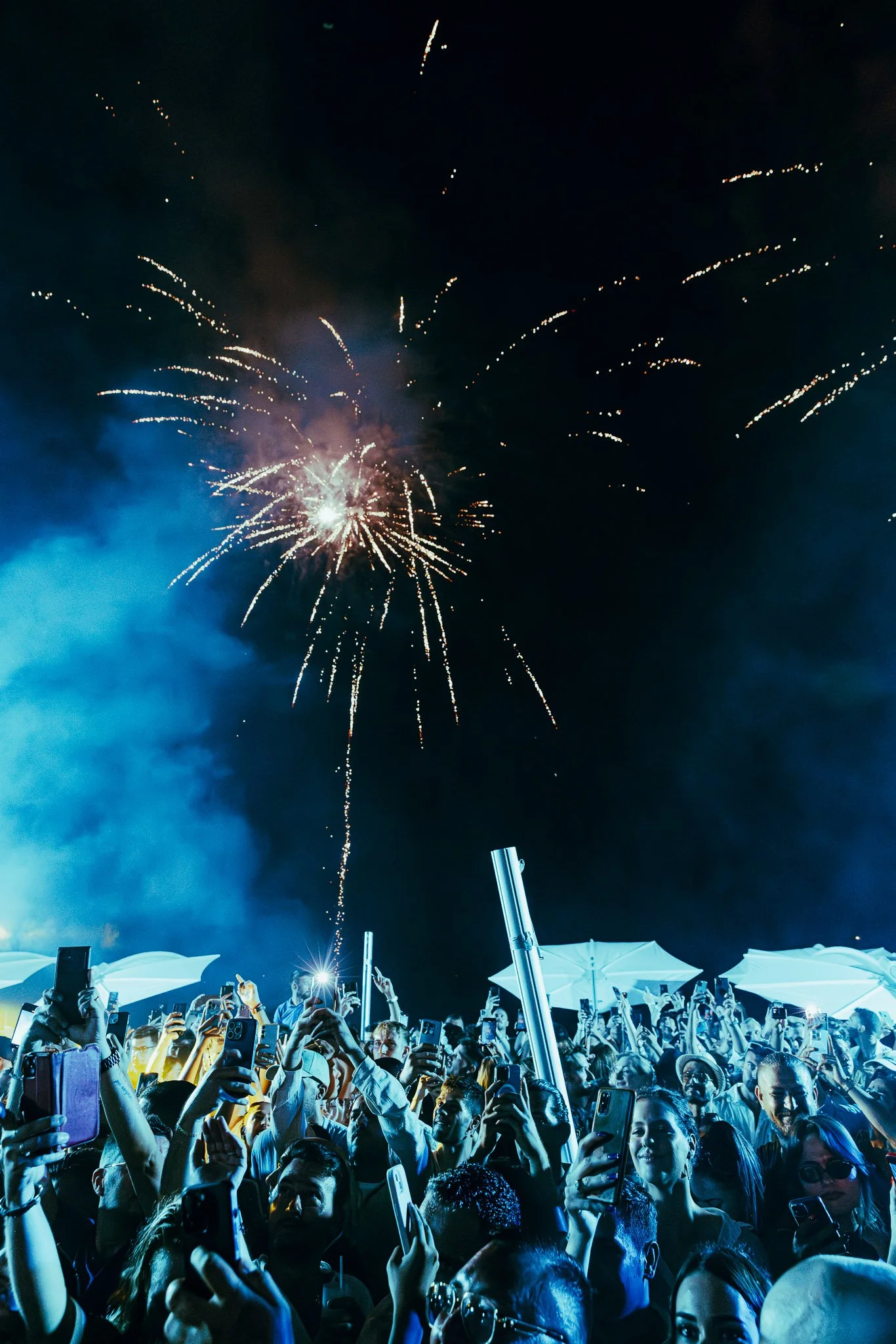 Crowd of people at night celebrating with fireworks in the sky, many holding up phones to record.