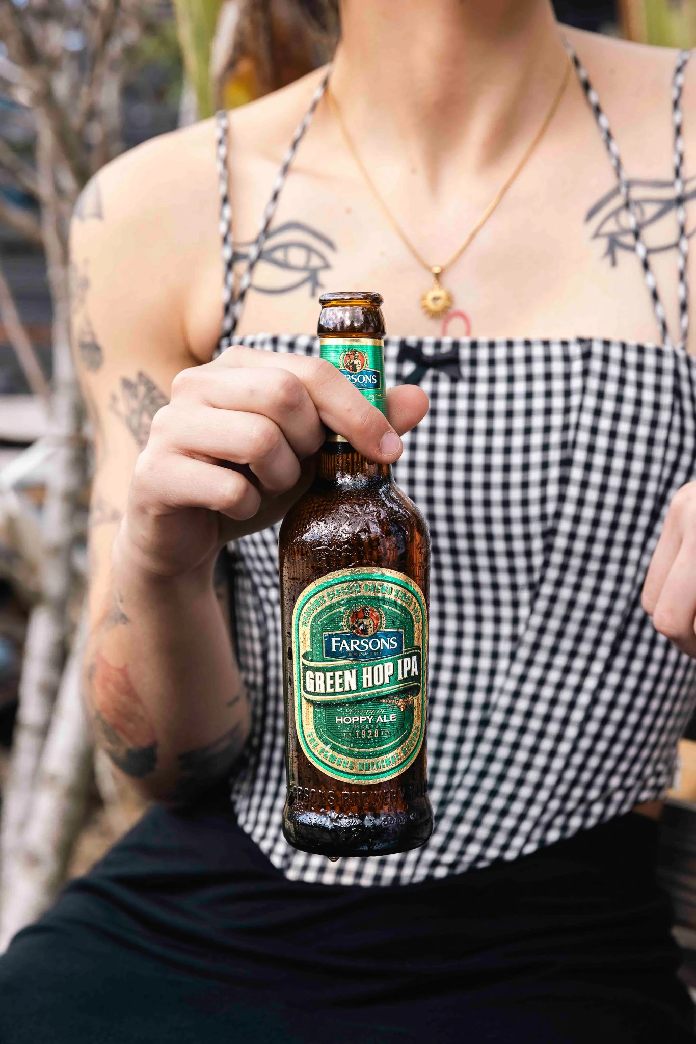 woman holding a green hop ipa bottle outdoors, wearing a plaid corset, tattoos, beer