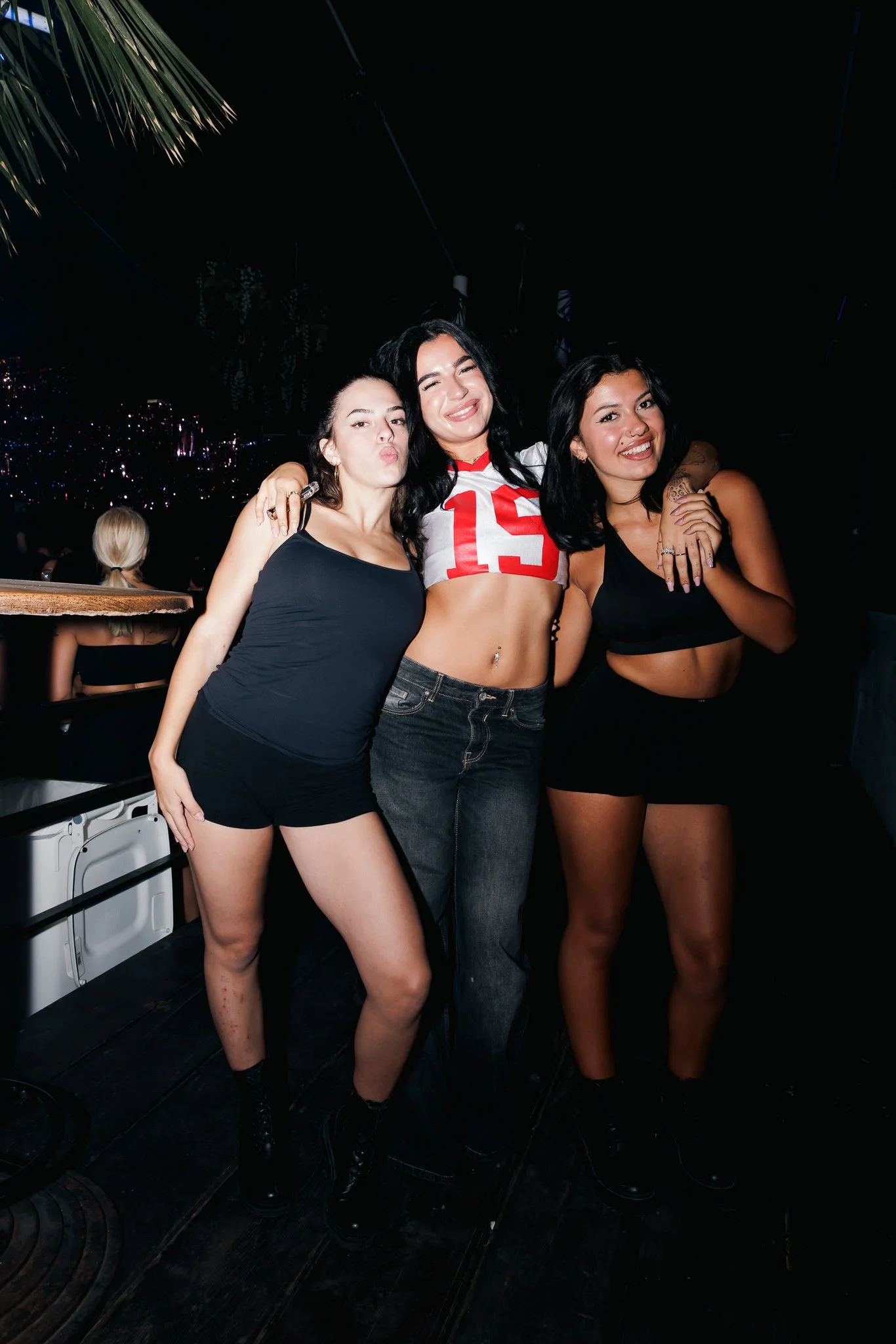 Three young women pose together in a nightclub, smiling and making playful expressions, with a dark background and some people in the distance.