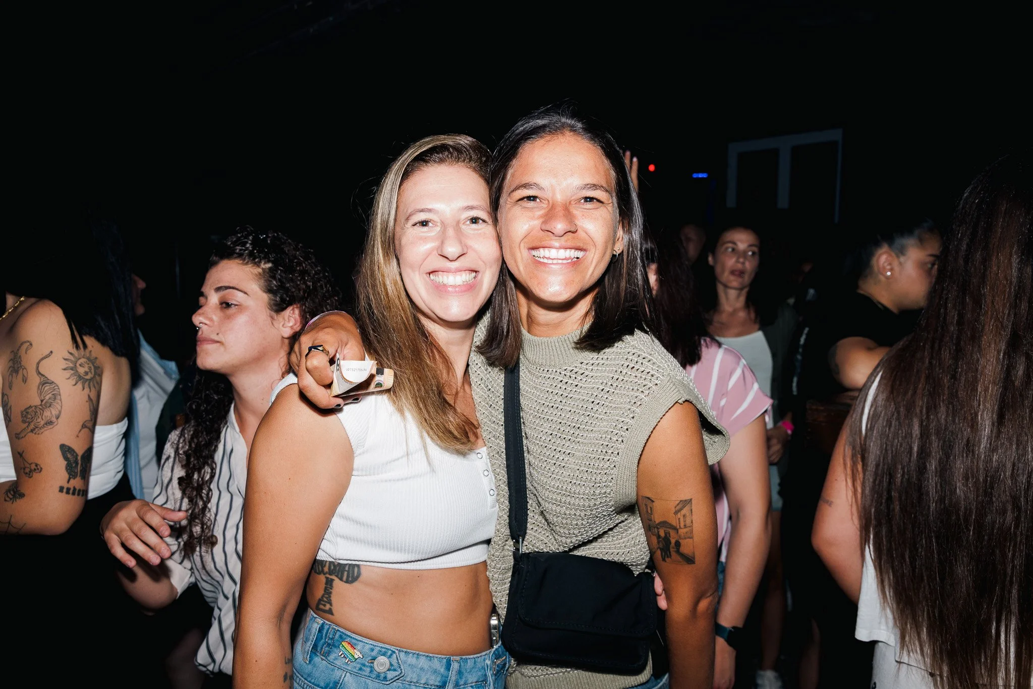 Two smiling women embracing at a crowded indoor event, with other people visible in the background.
