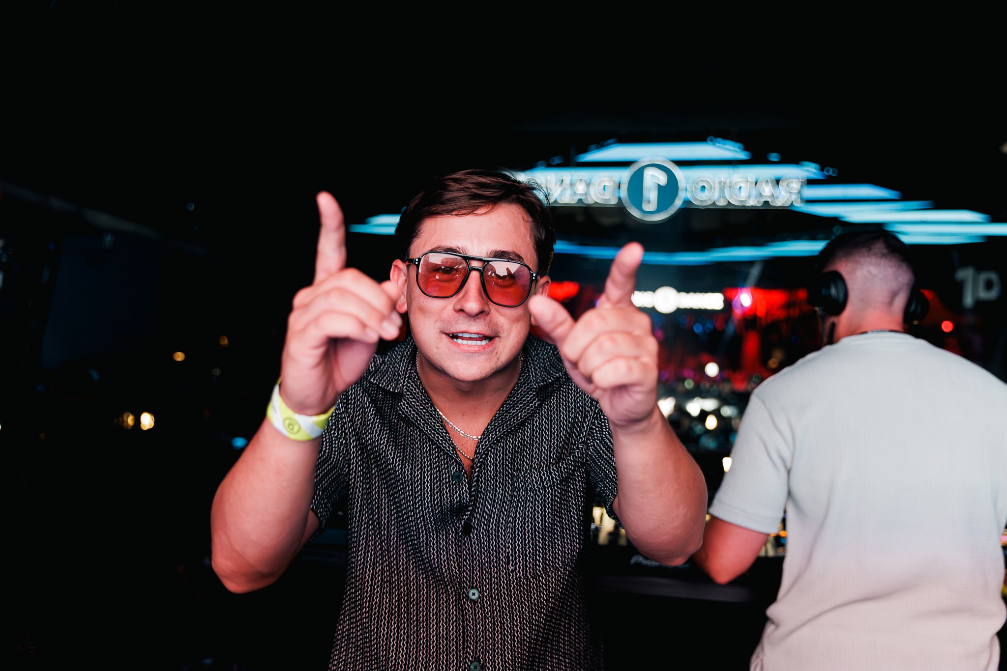 A man wearing sunglasses and a black patterned shirt making a gesture with both hands at a nightclub. There is a neon sign in the background that says 'PRADO DANCE' and another person wearing headphones is visible on the right.