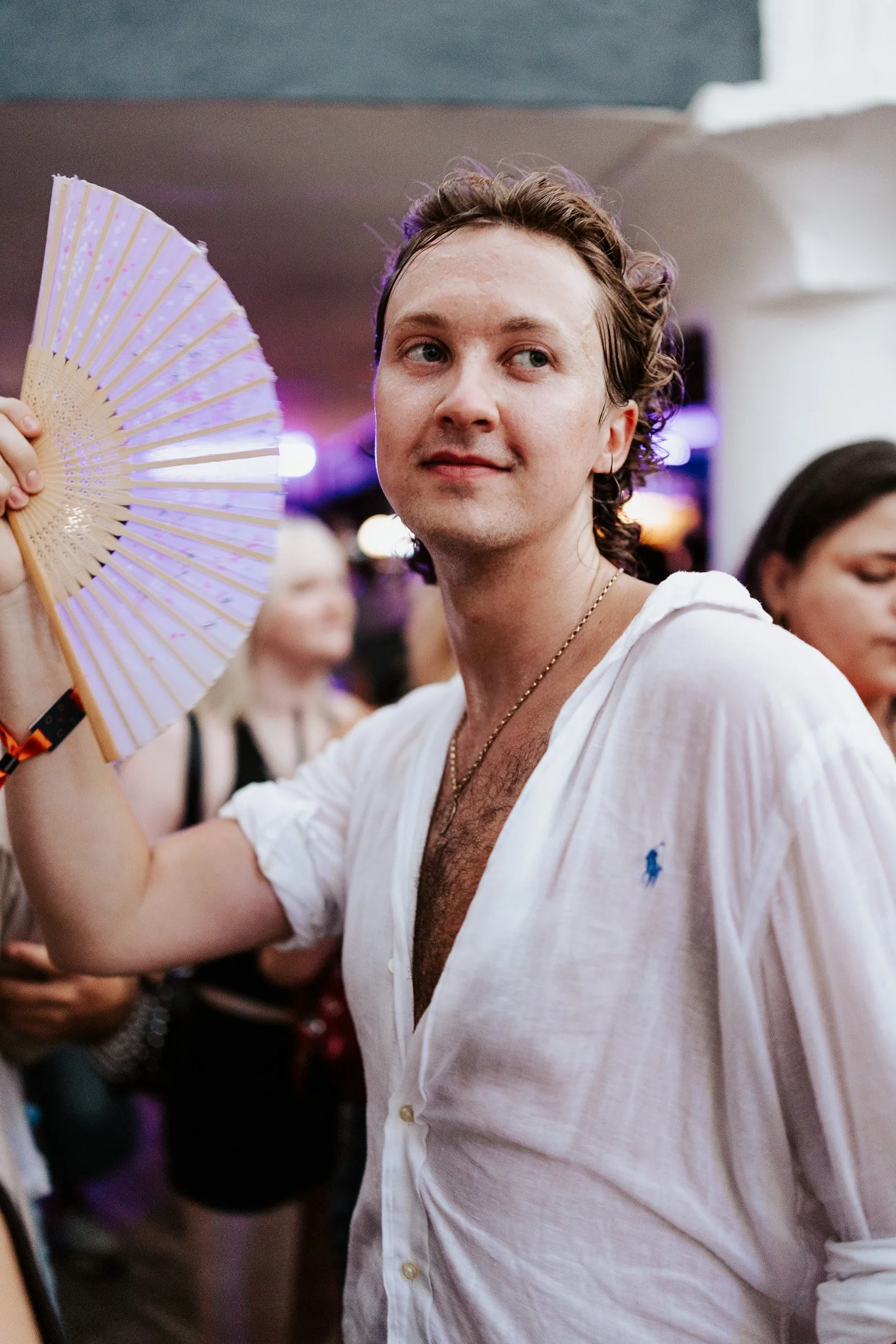 A man with curly hair and a necklace, wearing a white button-up shirt, holds a decorative fan near his face, smiling, in a setting with people in the background.