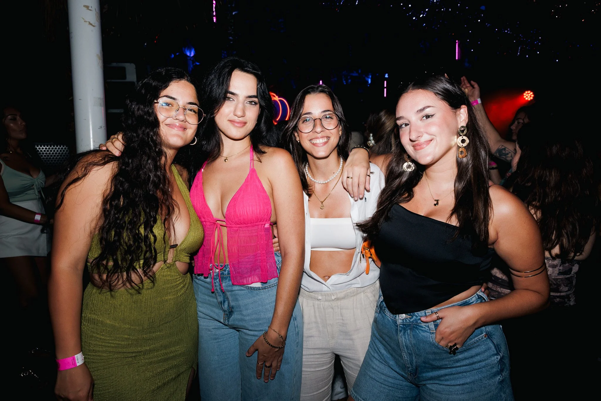 Four women posing together at a night event, smiling and dressed in colorful casual outfits, with a dark background and some purple and red lights.
