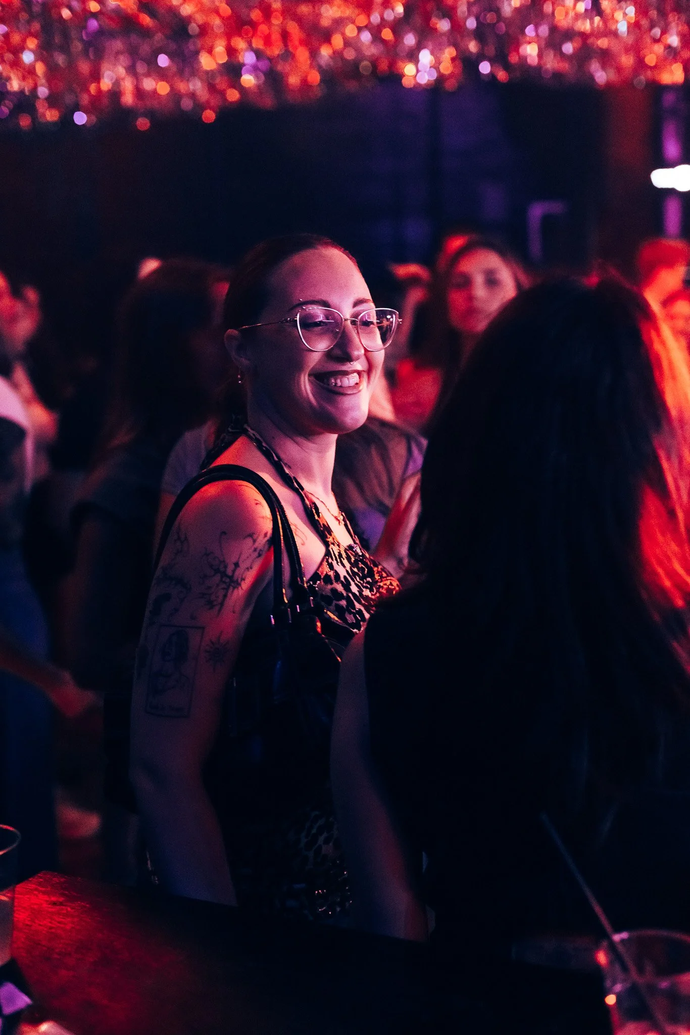 A woman with glasses smiling at a crowd at a concert or nightclub with colorful blurred lighting in the background.