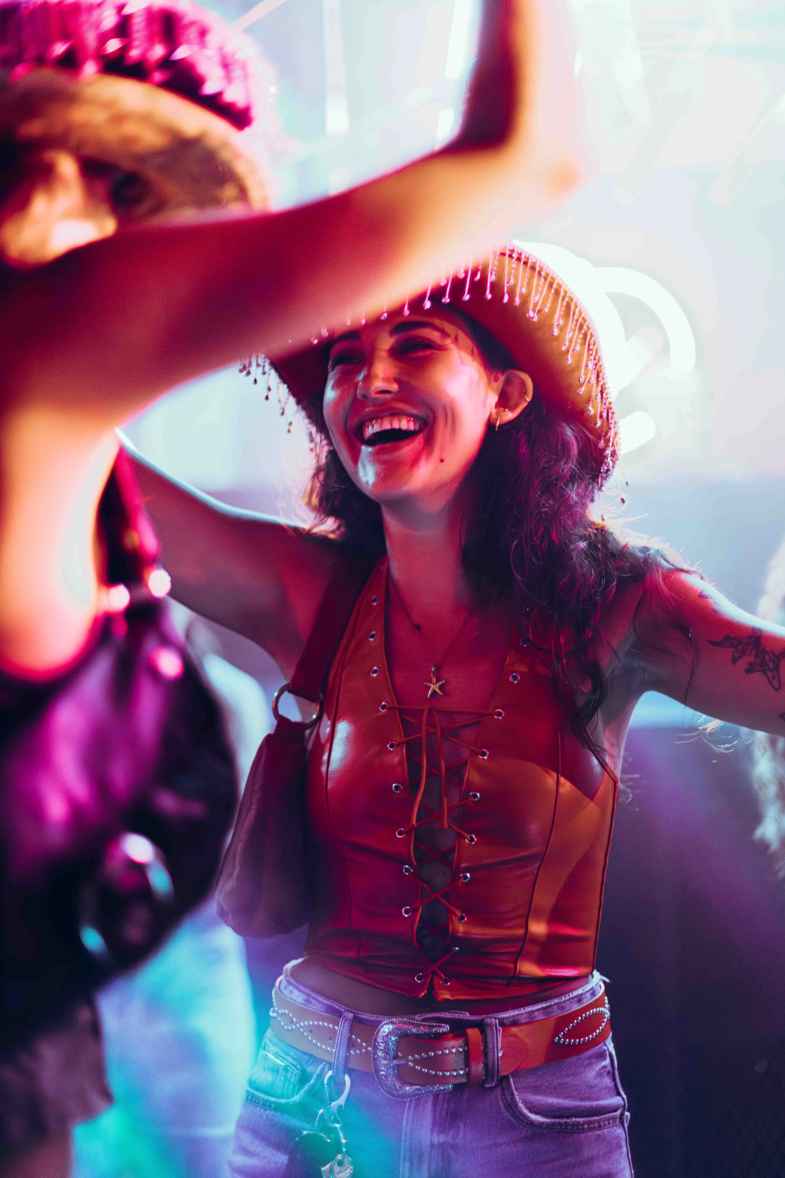 A woman wearing a sombrero hat and a red leather top is smiling and laughing while talking to someone. She is wearing earrings and a star necklace. The background has colorful neon lighting.