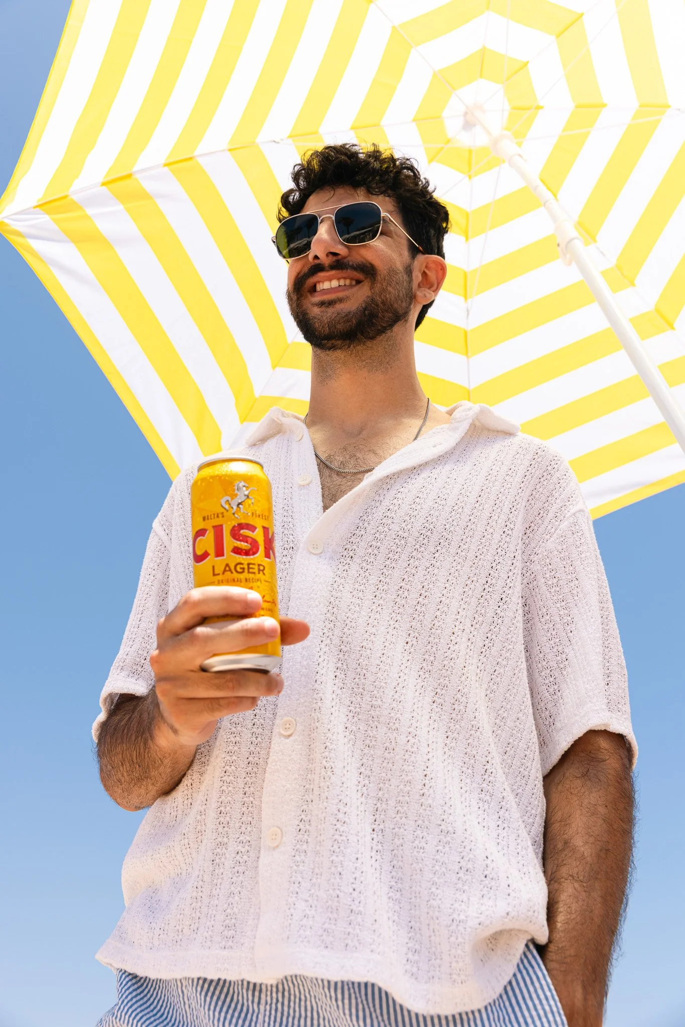 man by the beach, yellow striped umbrella behind him, smiling and holding a cisk lager can