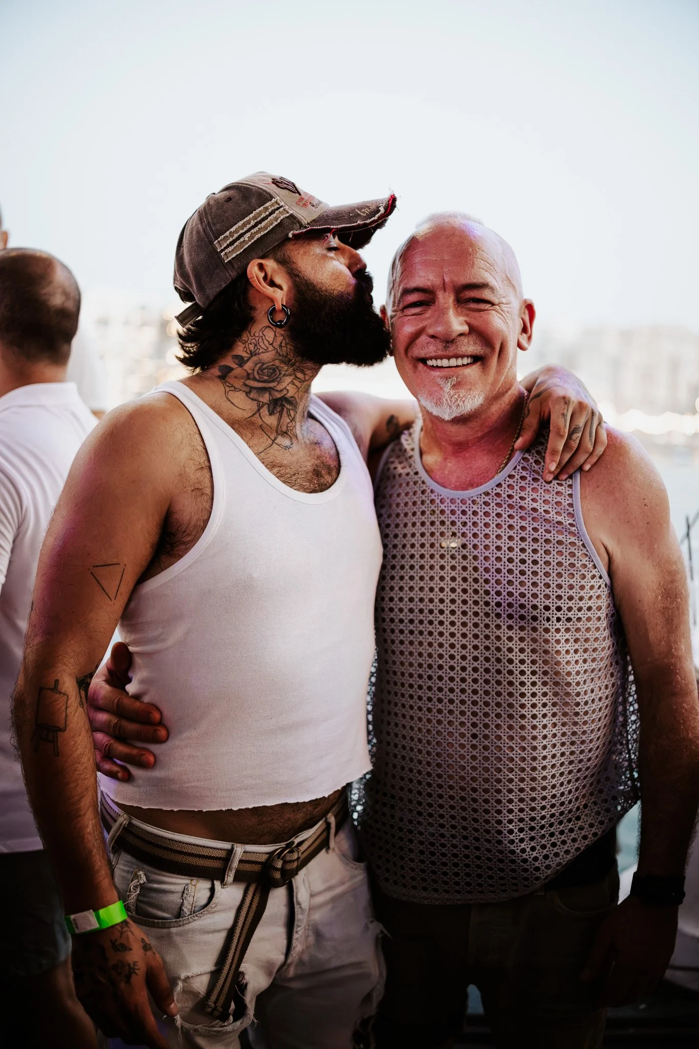 Two men are standing close together, smiling, with their arms around each other. One man has a beard, tattoos, and is wearing a brown cap and a white tank top. The other man is bald, smiling broadly, and wearing a mesh tank top. They are outdoors, wi