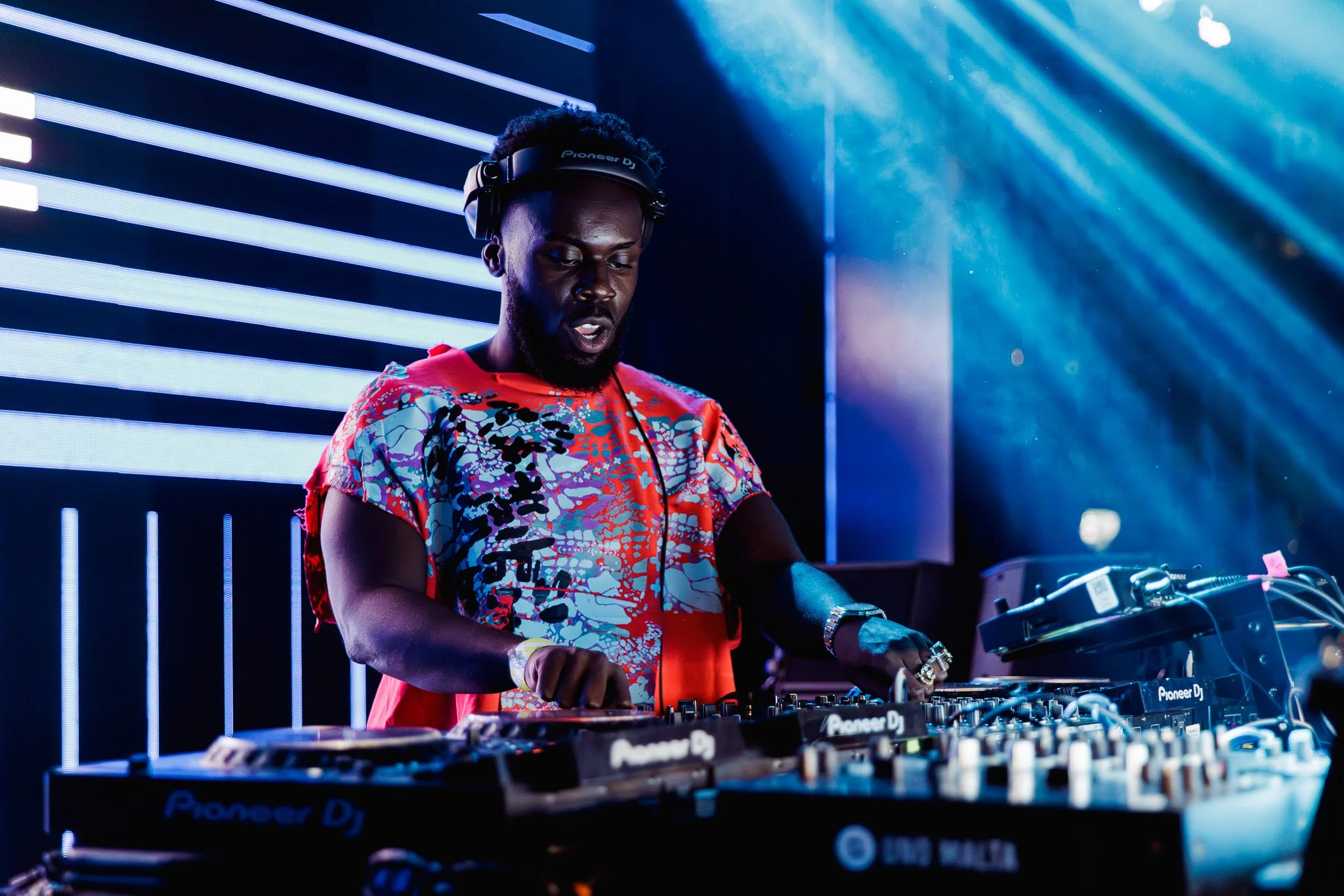 A DJ performing at a nightclub with neon blue lighting and modern decor. The DJ is wearing a colorful, patterned shirt, a Pioneer DJ headset, and is focused on the equipment.
