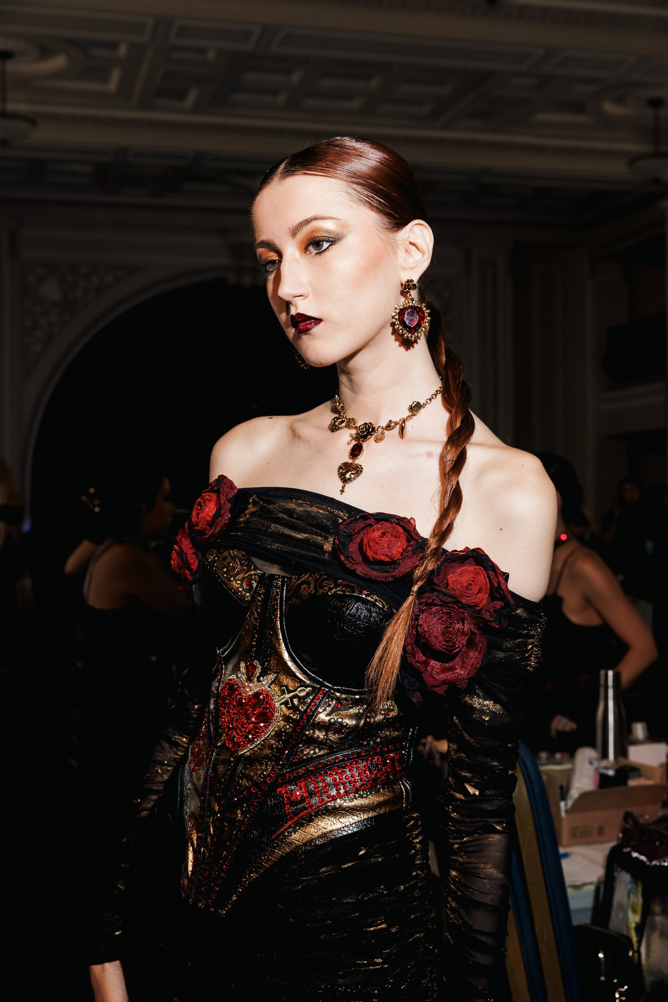 A woman with red hair in a braid wearing ornate jewelry and a dress adorned with red roses and gold accents, standing in an elegant interior space.