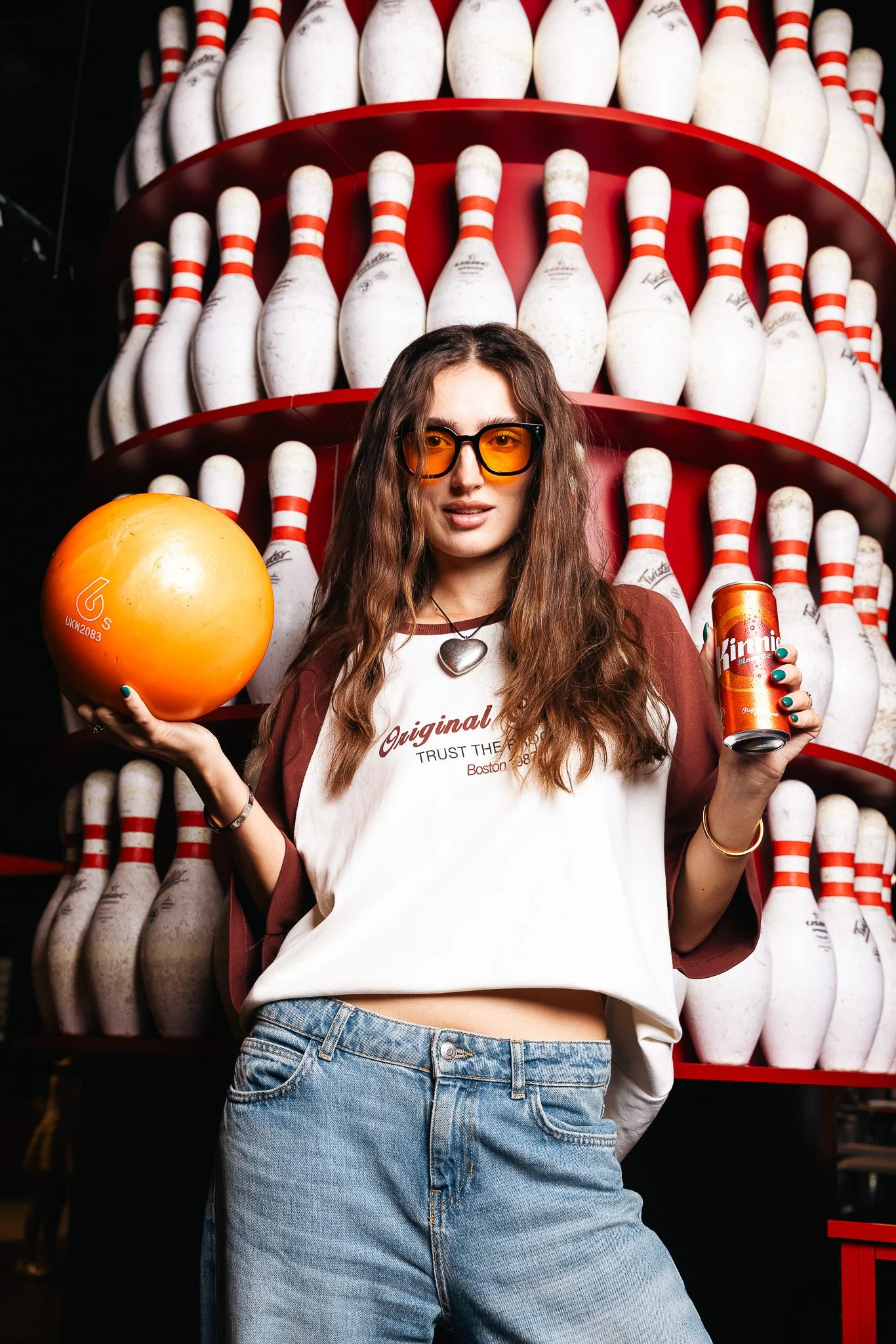 woman at a bowling alley, a wall of bowling pins behind her, holding a bowling ball and a kinnie can, cool pose with orange sunglasses