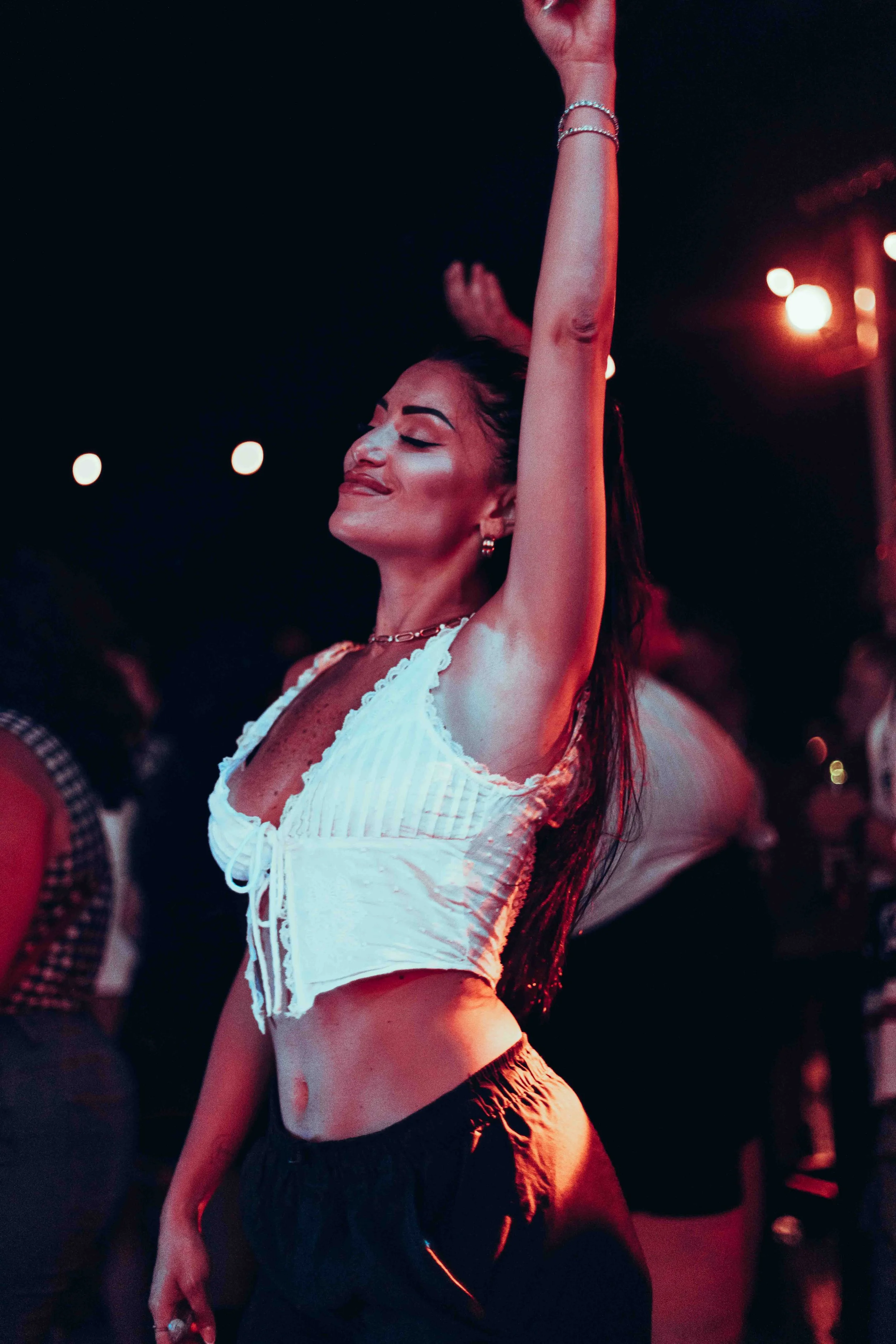 A woman dancing with her eyes closed at a lively event, wearing a white crop top and black shorts, with bracelets and earrings, under colorful stage lights.
