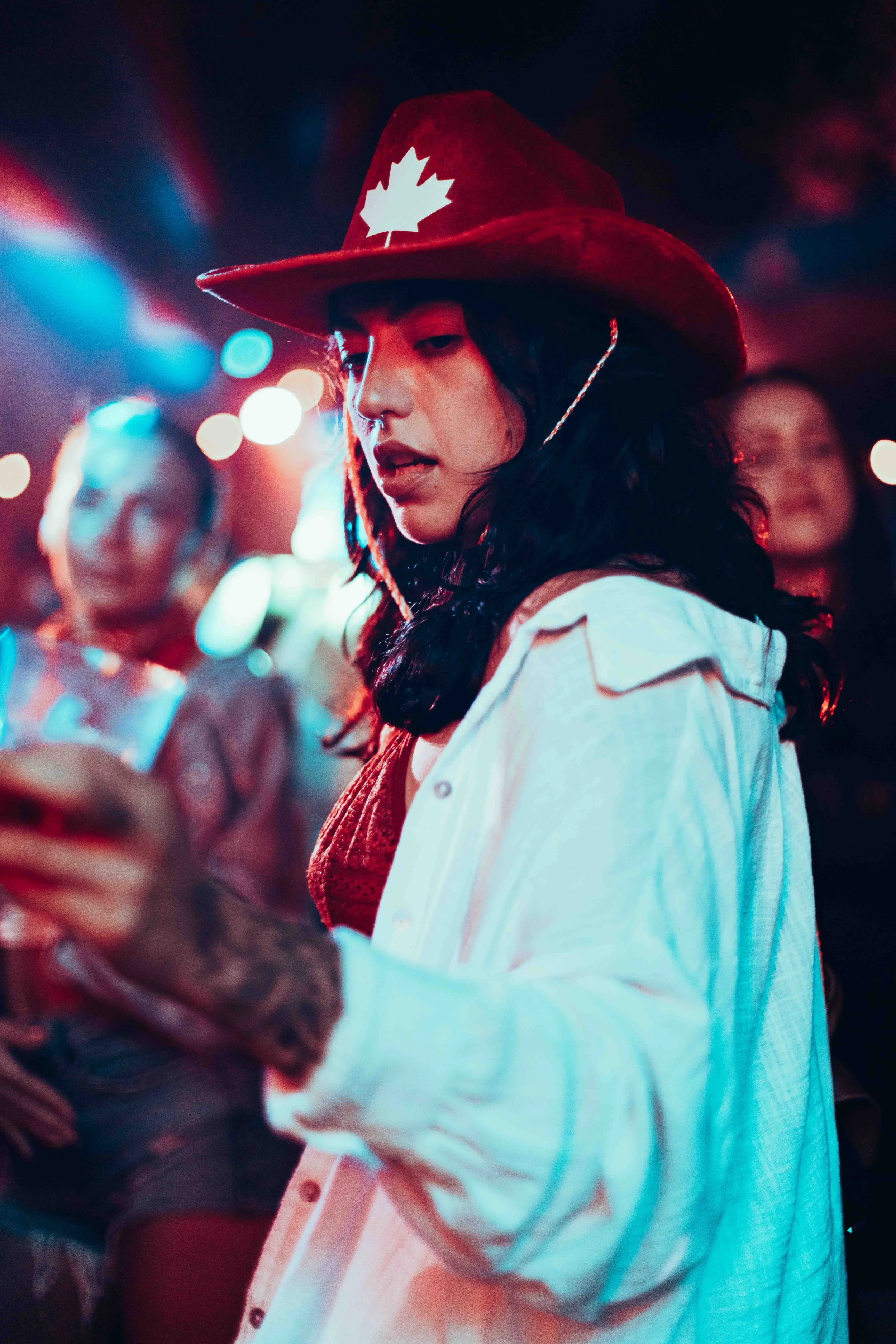 A woman wearing a red cowboy hat with a white maple leaf, black hair, and a nose piercing, standing in a dimly lit setting with colorful background lights, holding a smartphone.