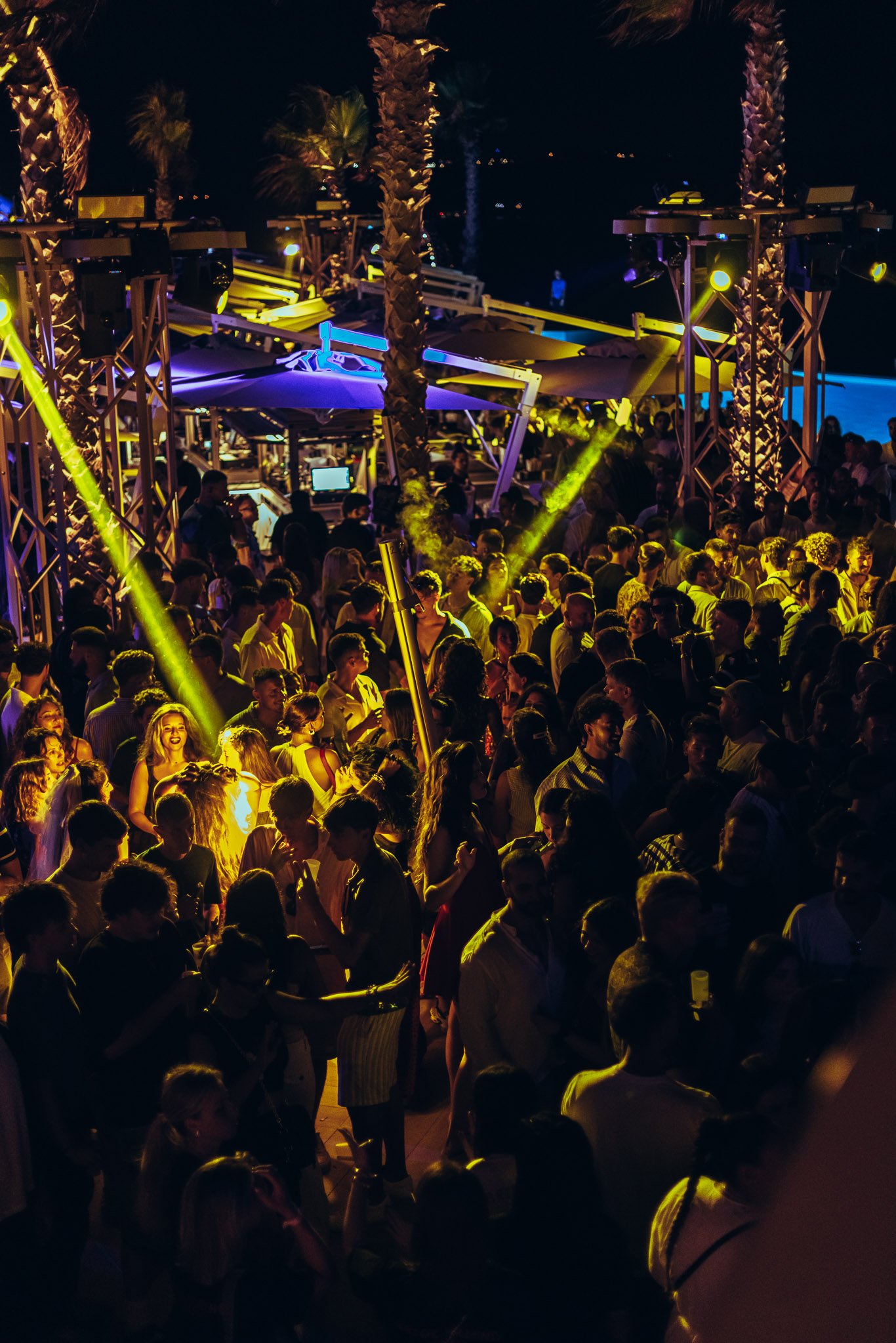A crowded outdoor nighttime party with many people dancing and socializing. Colorful stage lights and palm trees are visible, creating a lively atmosphere.