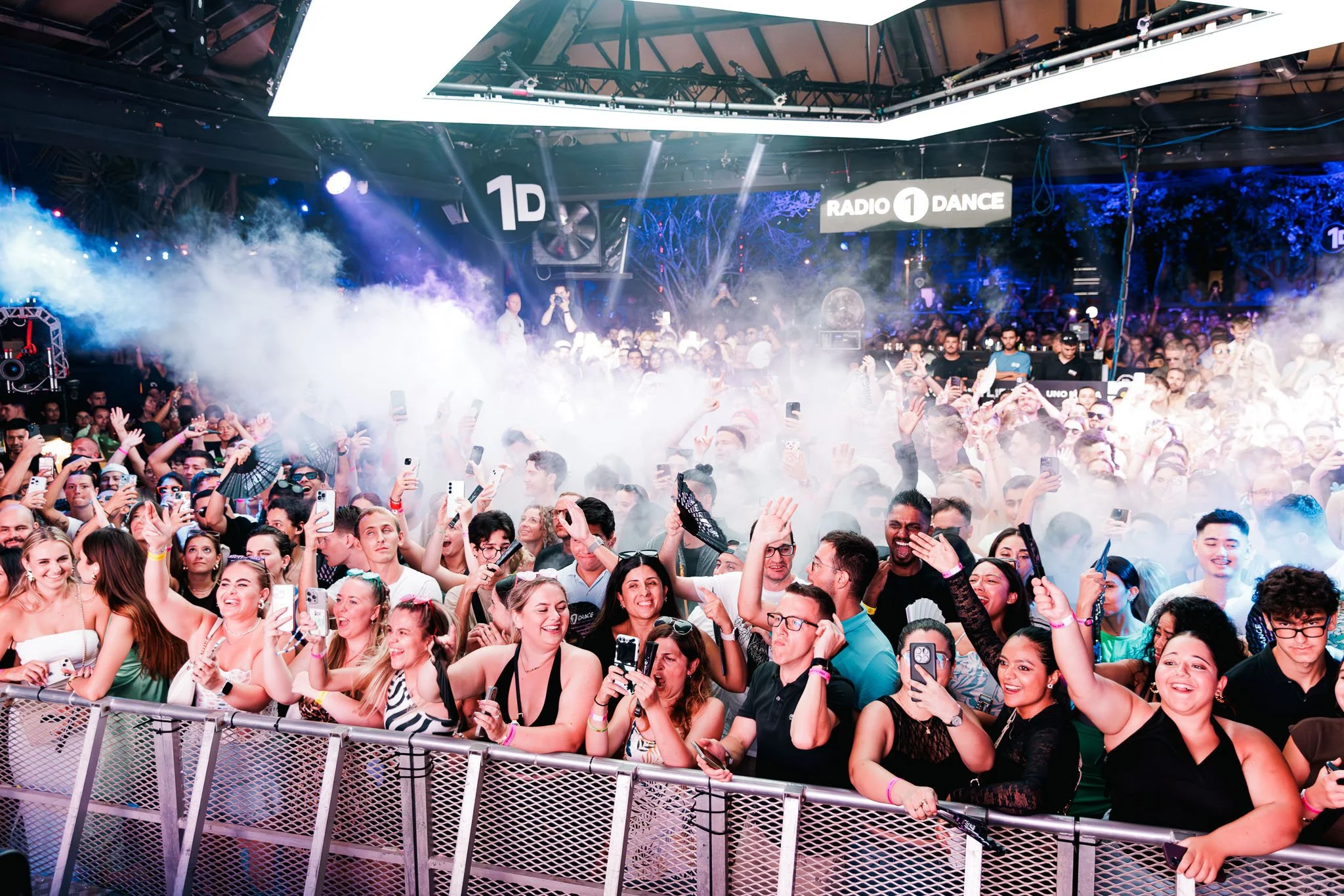Crowd at a concert or festival with many people smiling and holding phones, stage lights and smoke effects, and a sign that says 'Radio 1 Dance'.