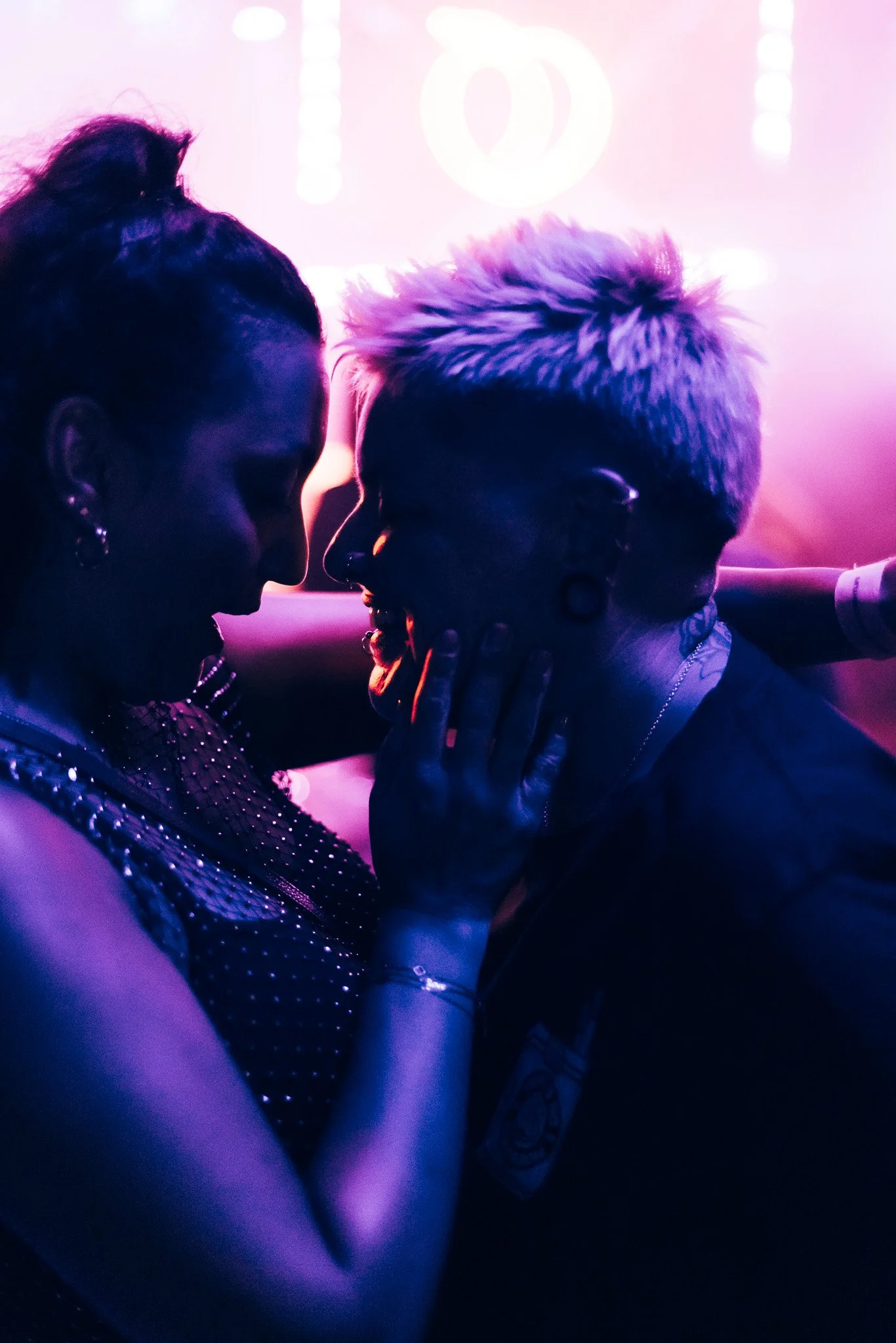 Two people are close together, facing each other in a dimly lit setting with purple and pink lighting. The person on the left has dark hair in an updo and is wearing a sparkly outfit. The person on the right has short, styled hair and is touching the