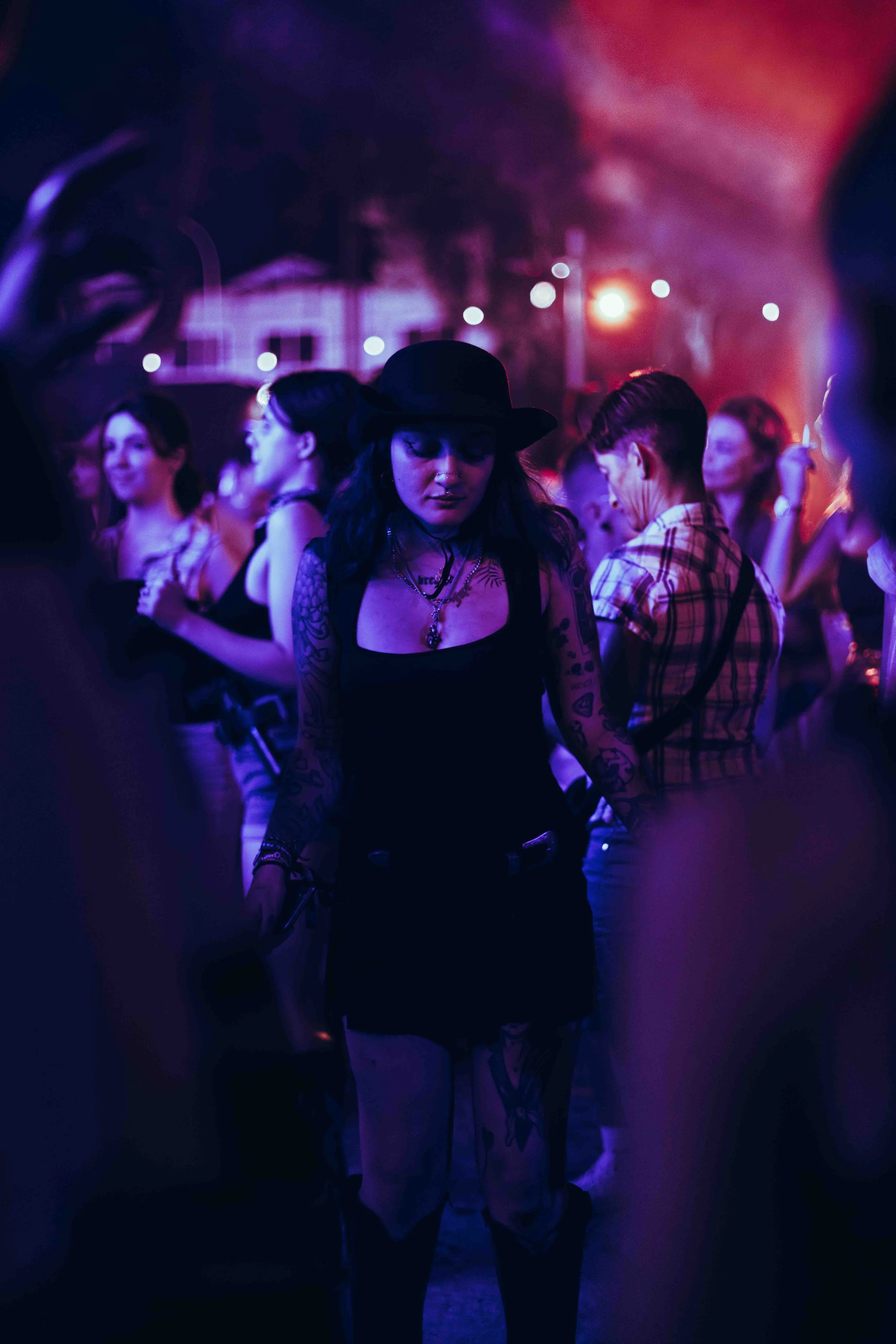 A group of people at a nighttime party or concert with purple and red lighting. A woman in the foreground wears a black hat, has dark hair, tattoos on her arms, and is dressed in dark clothing. Other women are visible in the background, engaged in co