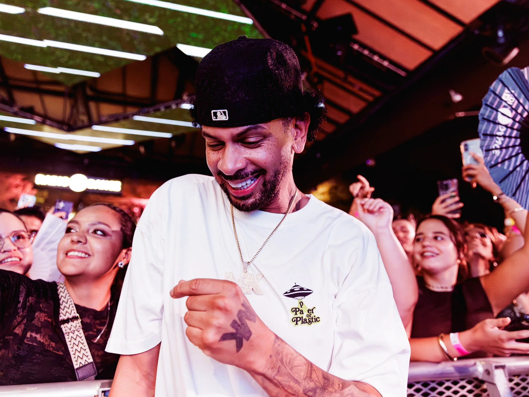 A man with a beard wearing a black baseball cap with a Major League Baseball logo, smiling and holding hands with a woman. The man is wearing a white T-shirt with a logo that reads 'Paper or Plastic' and a UFO image. They are surrounded by a crowd of