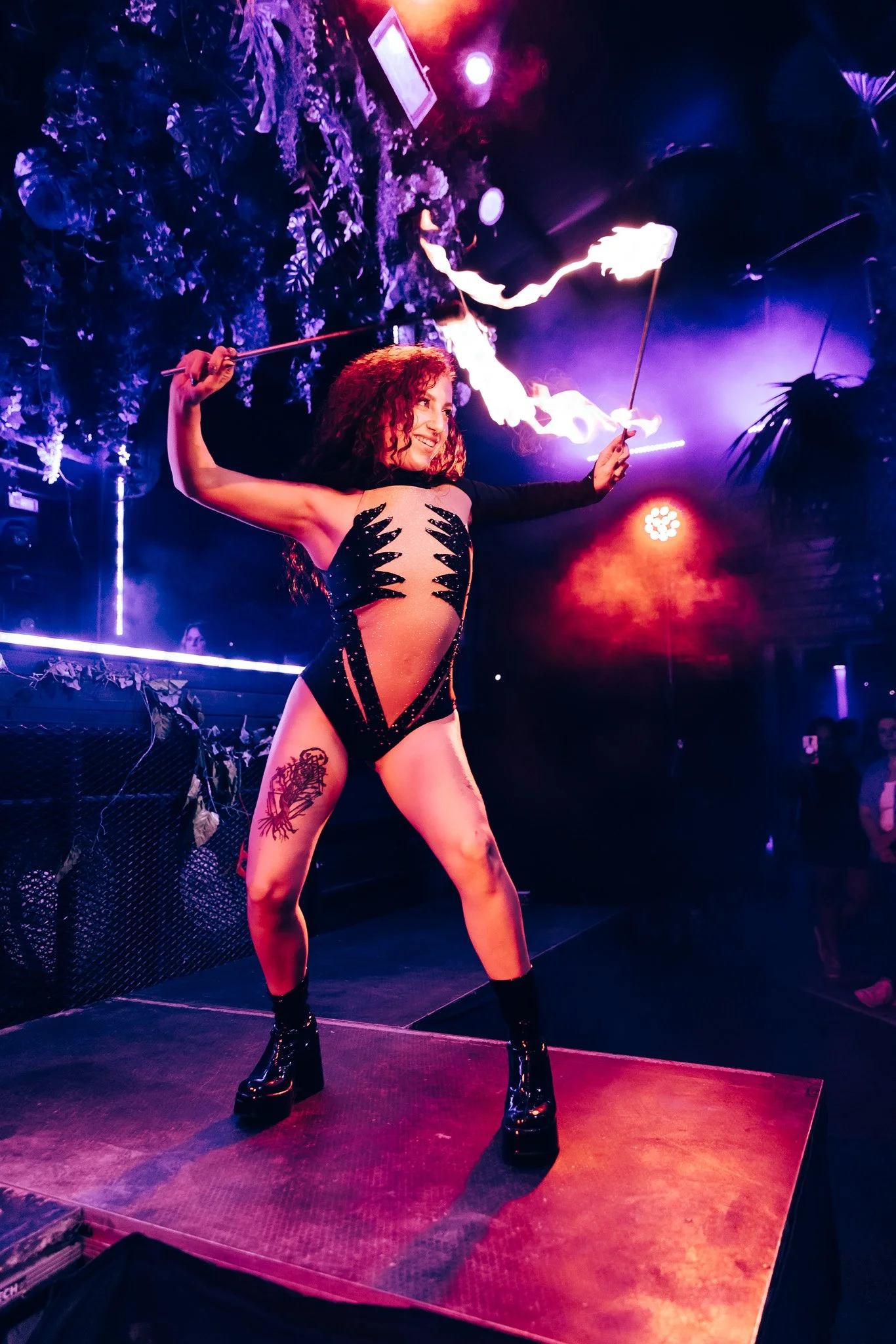 A woman with curly red hair performs a fire dance on a stage with purple lighting, plants, and dark background.