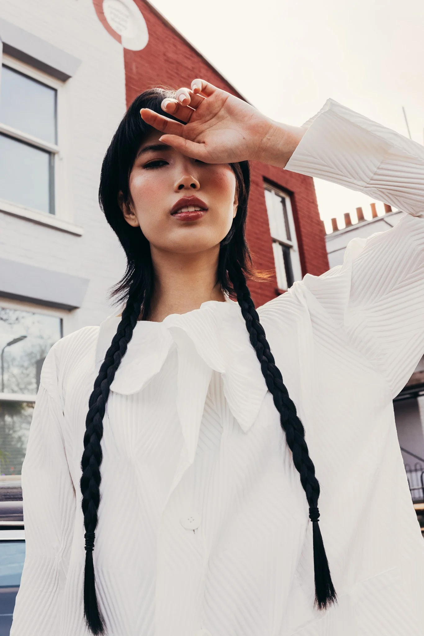 A woman with black hair styled in two braids standing outdoors, shielding her eyes from the sun with her right hand, with modern buildings in the background.