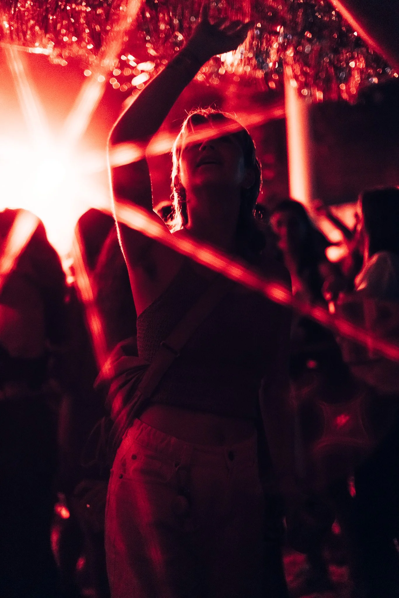 Person with long hair taking a selfie in a dimly lit space with red lighting, surrounded by other people and reflections.