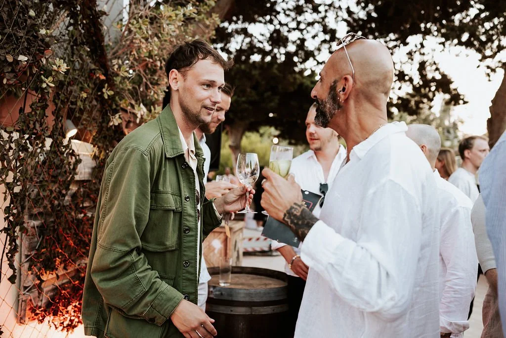 Two men are talking and holding drinks at an outdoor gathering, with other people in the background.