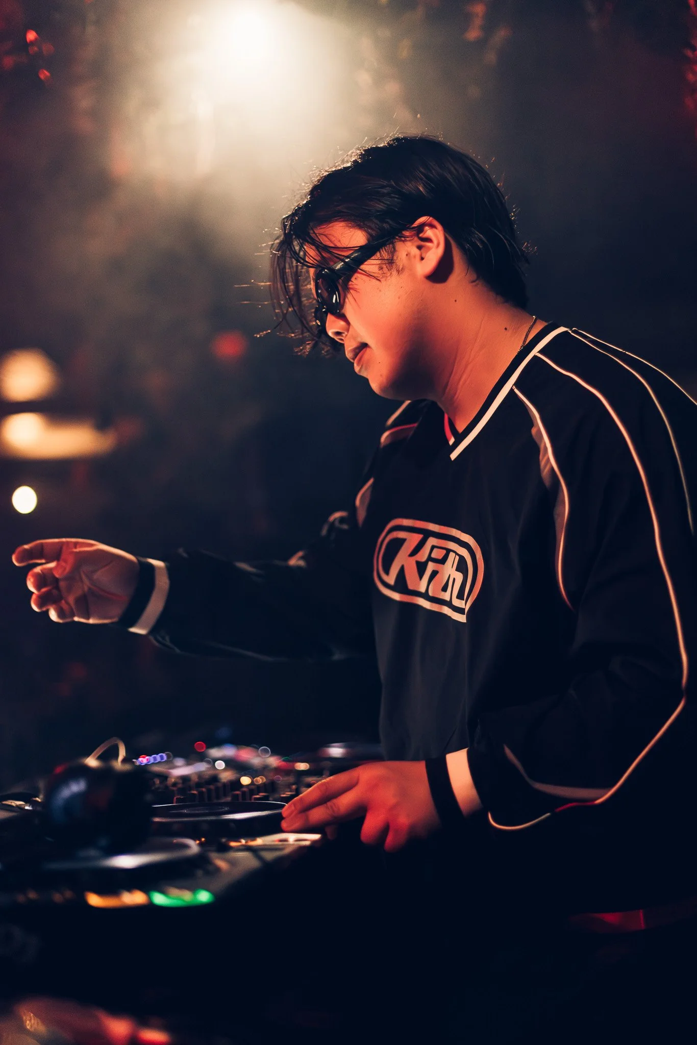 A DJ wearing sunglasses and a black racing jacket with white striping and logo, mixing music in a dimly lit club with warm lighting and blurred colorful lights in the background.