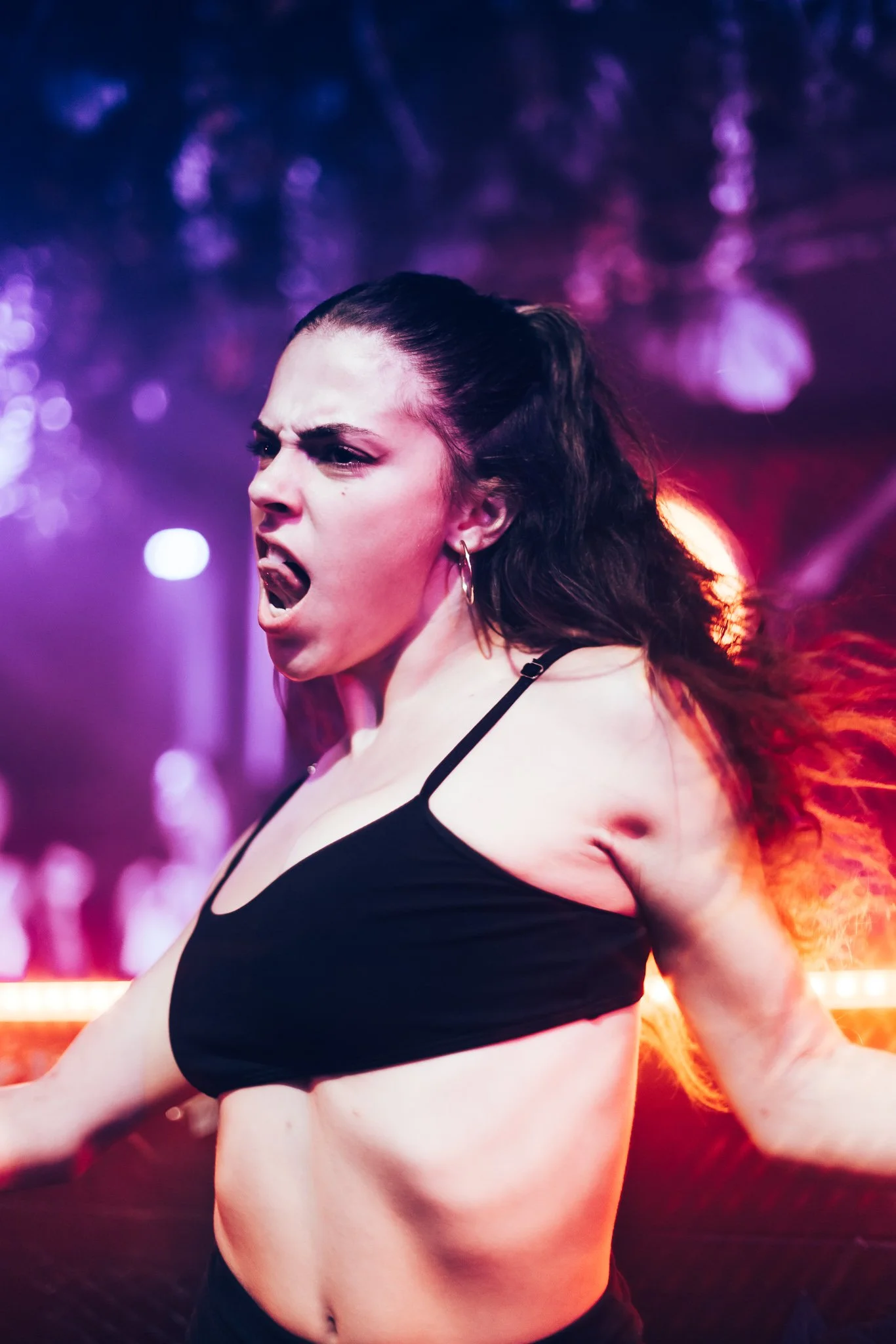 A woman with dark hair in a ponytail, wearing a black sports bra, appears to be dancing or performing energetically against a colorful, vibrant light background with purple, pink, and orange hues.
