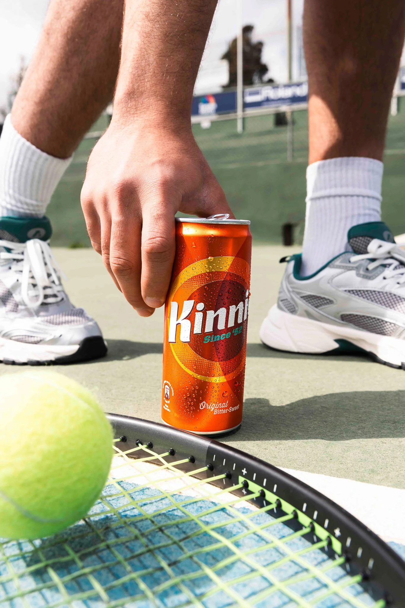 kinnie can next to a tennis racket and tennis ball, person reaching for it, social media shoot