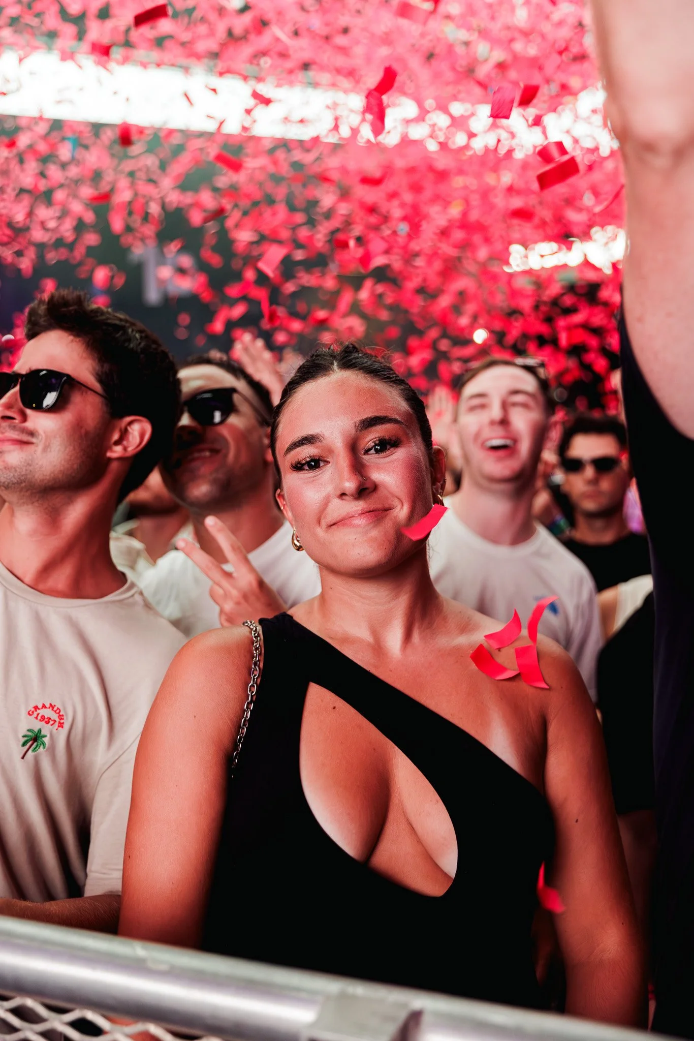 A group of people celebrating with red confetti falling from above. The woman in the foreground is smiling and making a peace sign with her fingers, wearing a black dress with a strap. There are several men in the background, some wearing sunglasses,