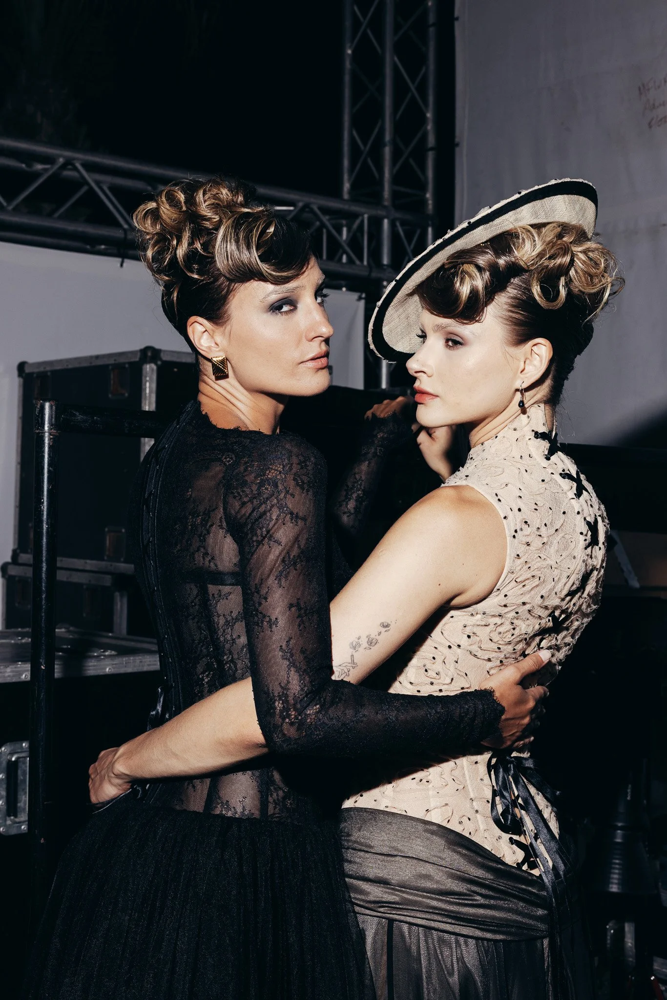 Two women with elaborate hairstyles and vintage-inspired fashion, standing very close and embracing each other backstage at an event.