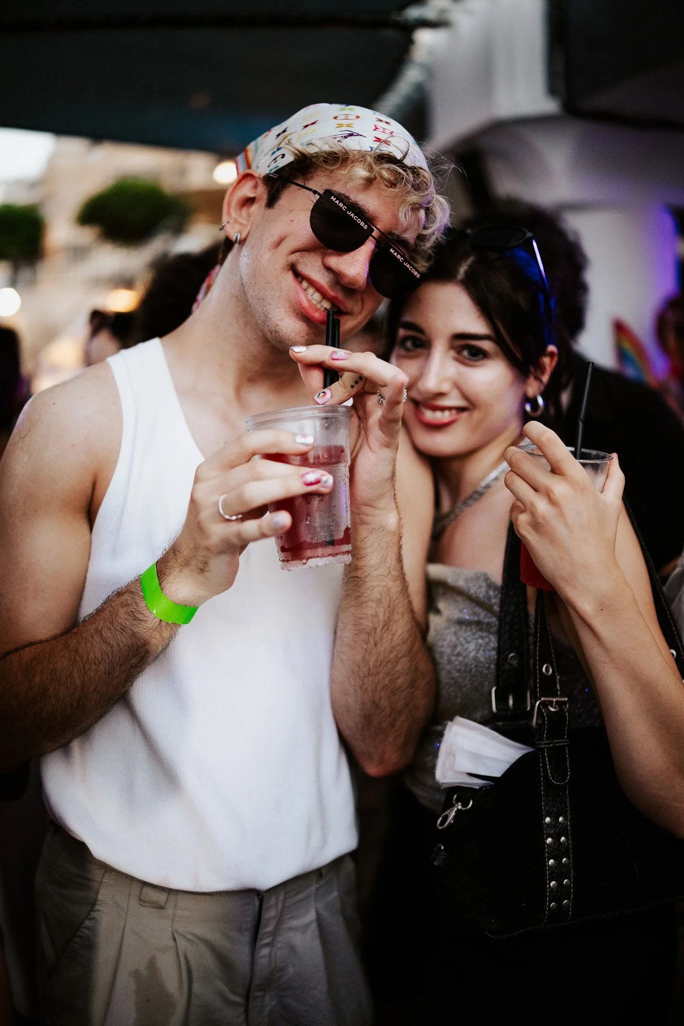 A man and woman smiling and holding drinks at a party or social gathering, with the man wearing sunglasses and a bandana, and the woman wearing glasses and black clothing.