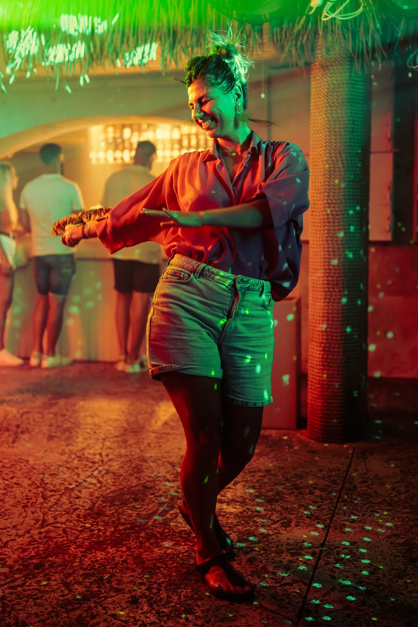 A woman dancing at a nightclub with green and red lighting, wearing a red satin shirt, denim shorts, and sandals, smiling with her hair in a messy bun, with other people in the background.