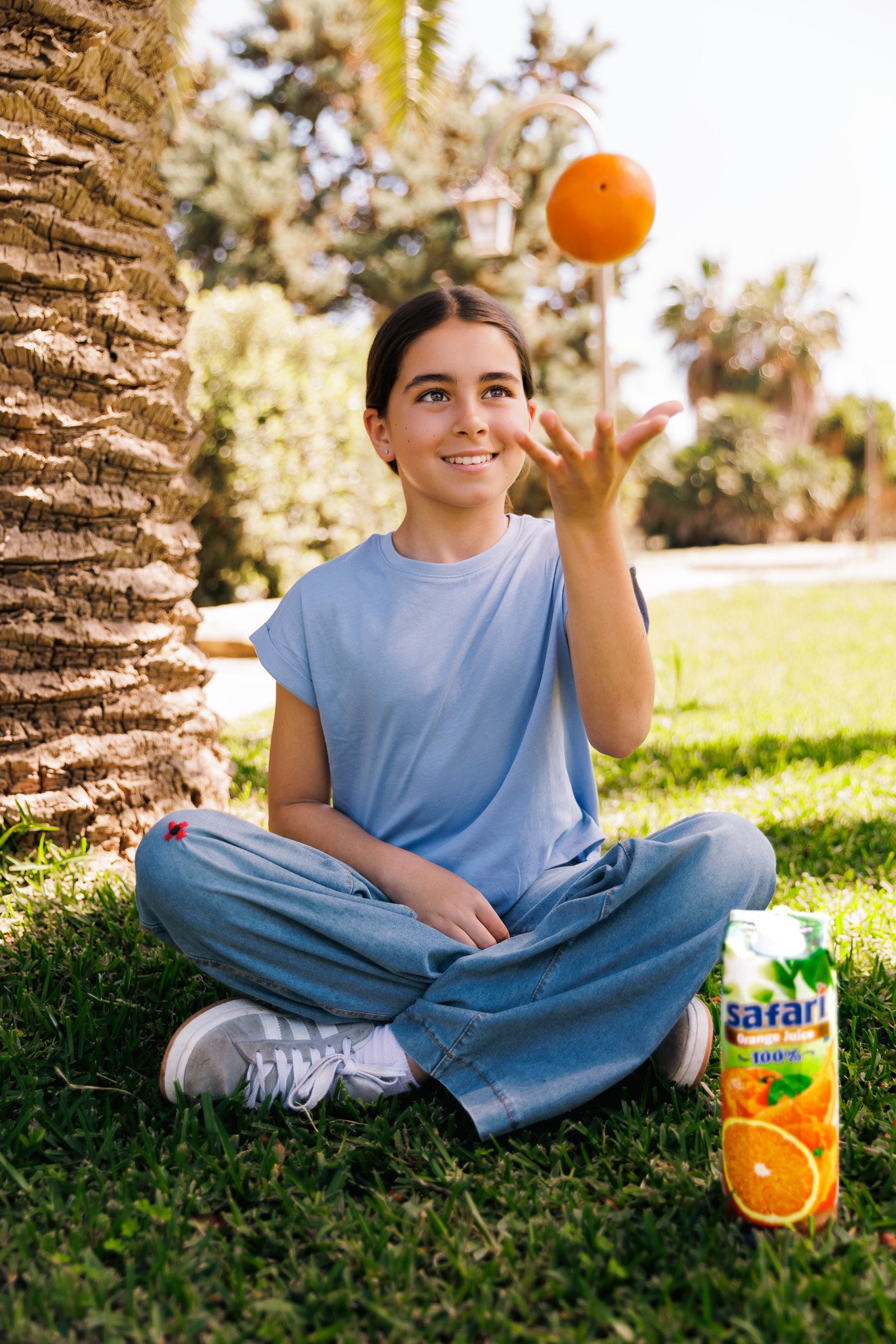 young girl throwing an orange in the air, sitting down on the grass, safari juice box next to her, commercial photoshoot